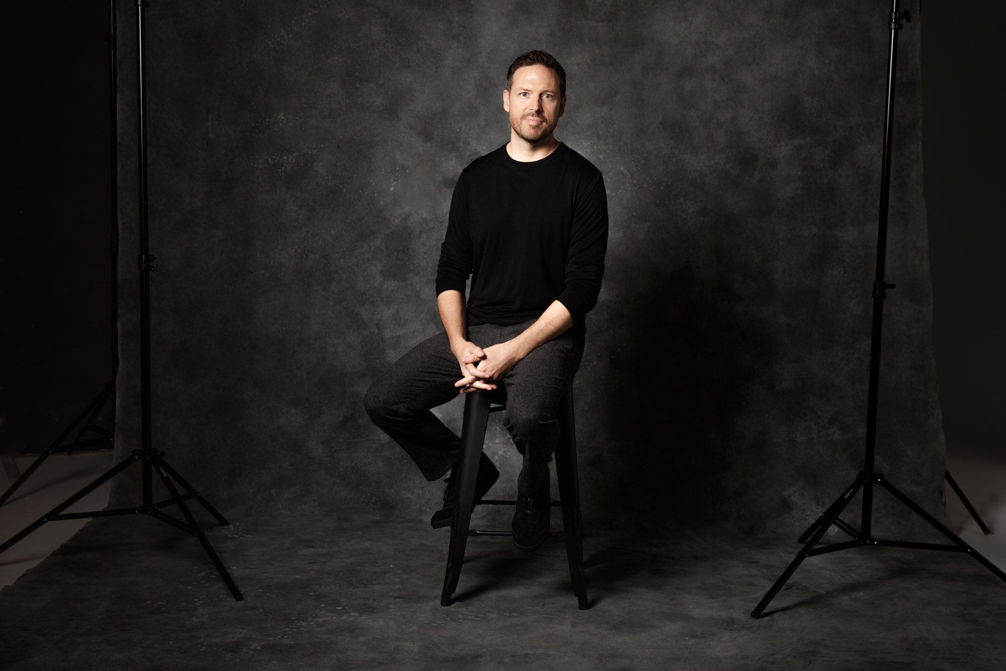 A man with a beard and mustache sitting on a stool in a photography studio with a dark textured backdrop, flanked by studio lights.