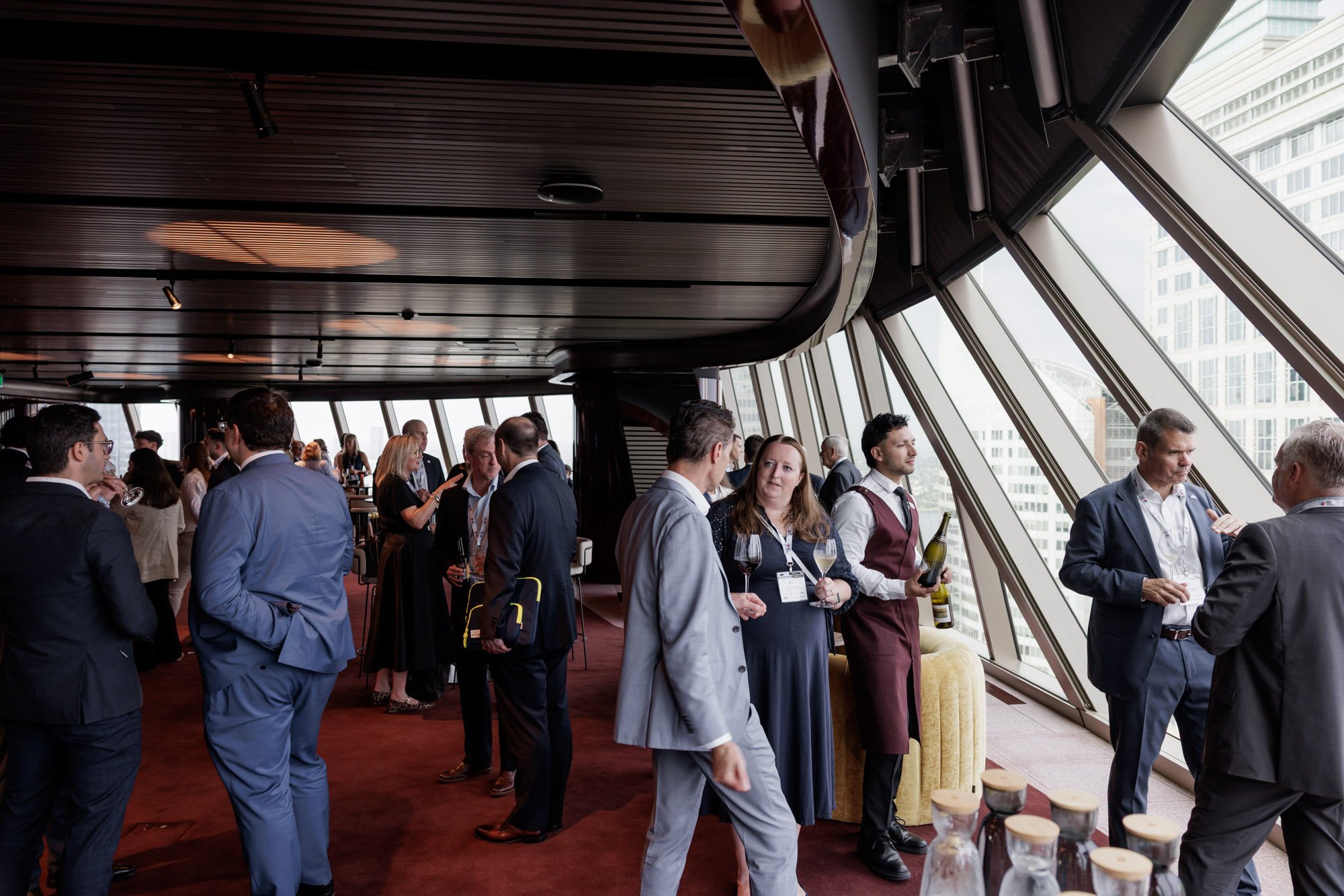 Business professionals socializing and networking at a corporate event in a modern, high-rise building with large windows and city views. Some hold glasses of wine, while others have name tags.