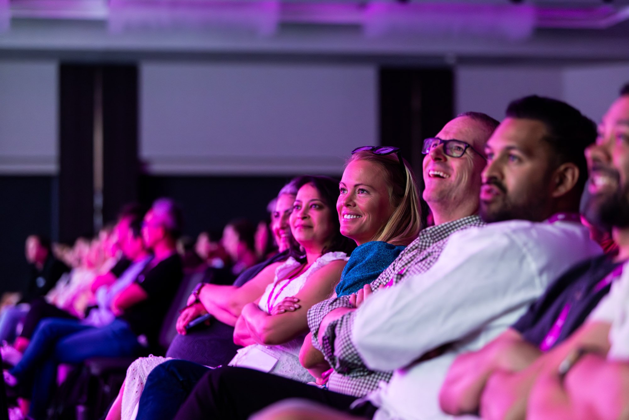 Audience watching a presentation or performance, illuminated with purple and pink lighting.