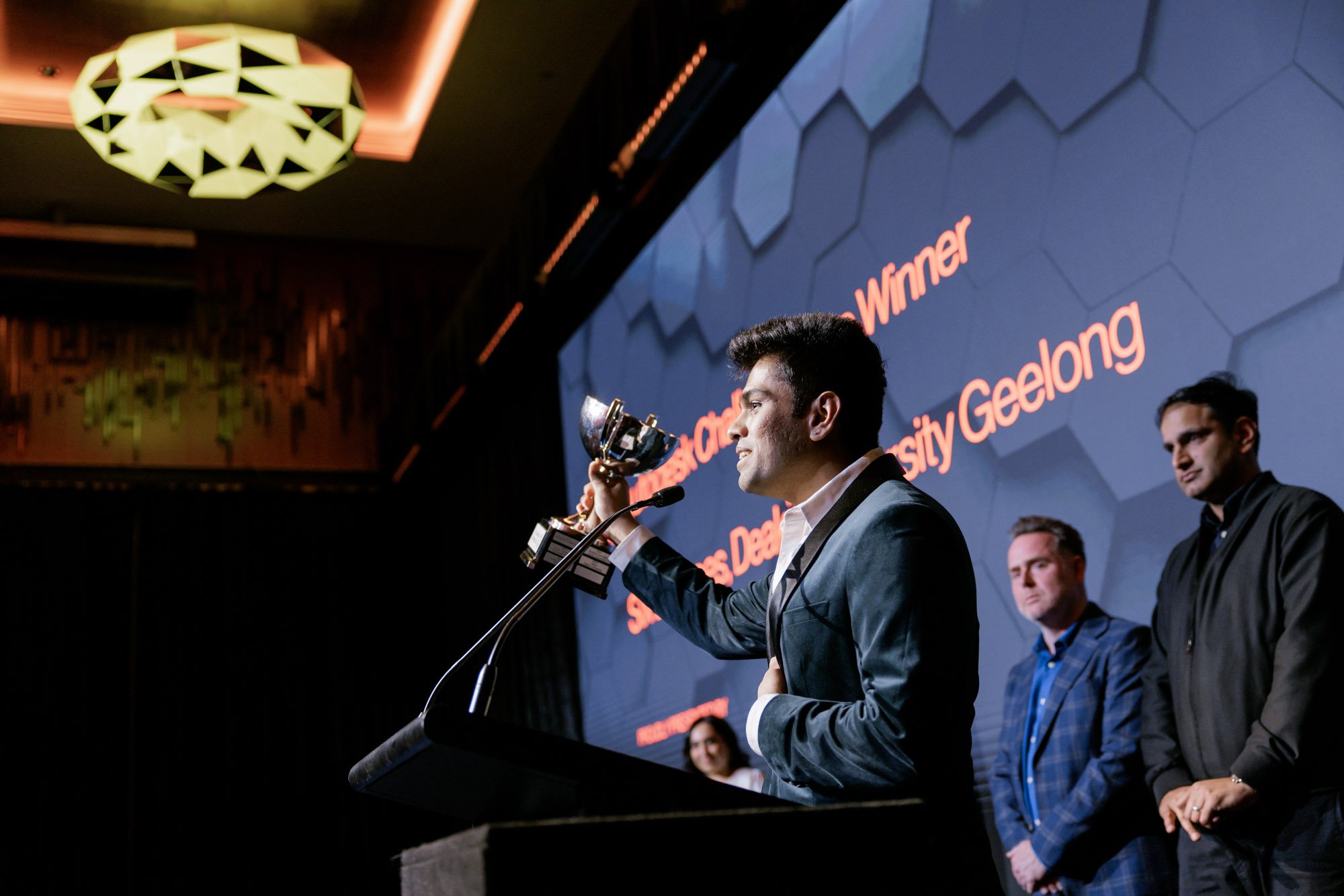 A man in a suit holding a trophy while speaking at a podium during an awards ceremony, with a large screen behind him displaying the words 'Winner' and 'Sustainable Development Goals'.