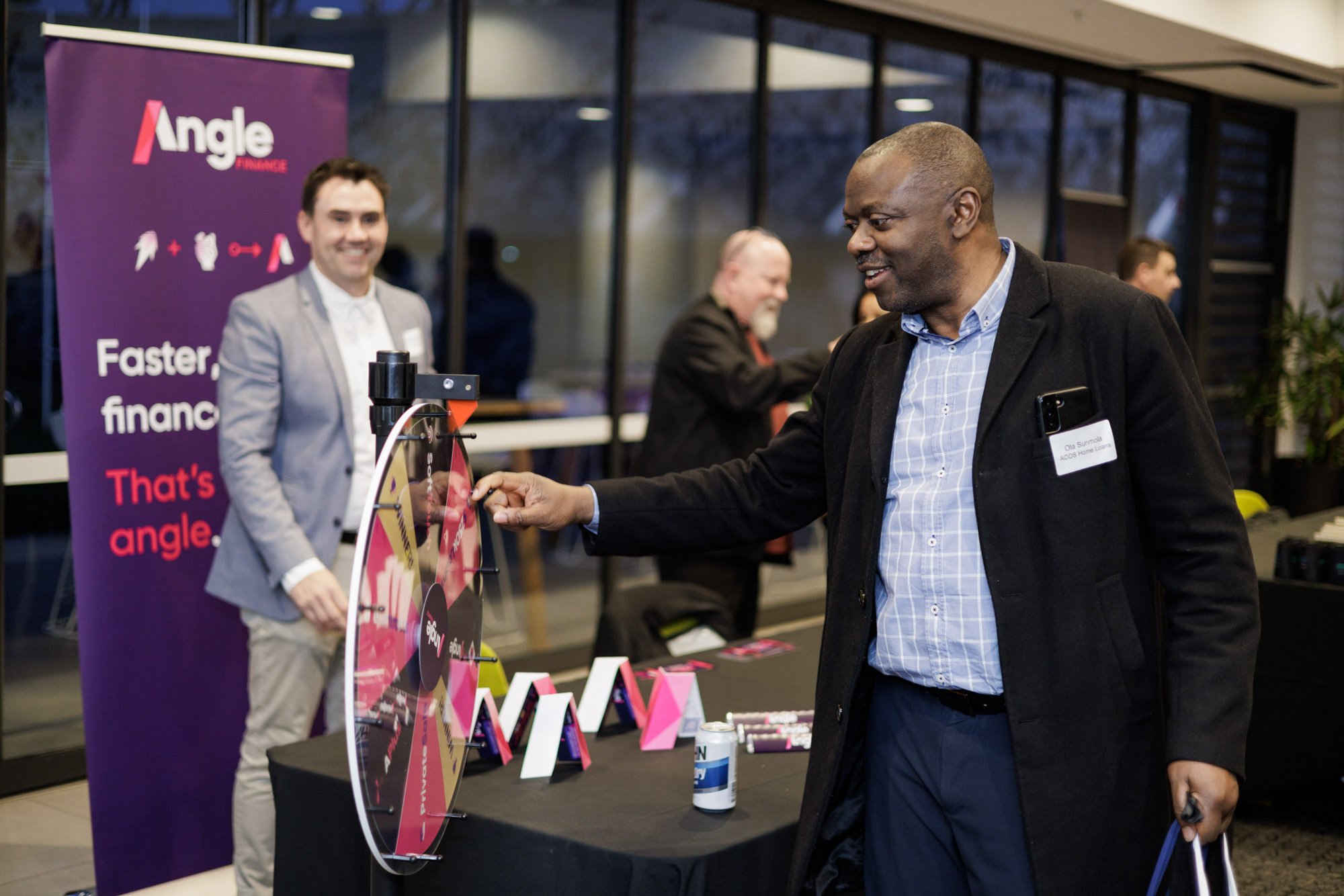 A man in a black coat and blue shirt playing a spin the wheel game at a business event, with a purple Angle Finance banner in the background. Other people are visible behind him, engaging in the event.
