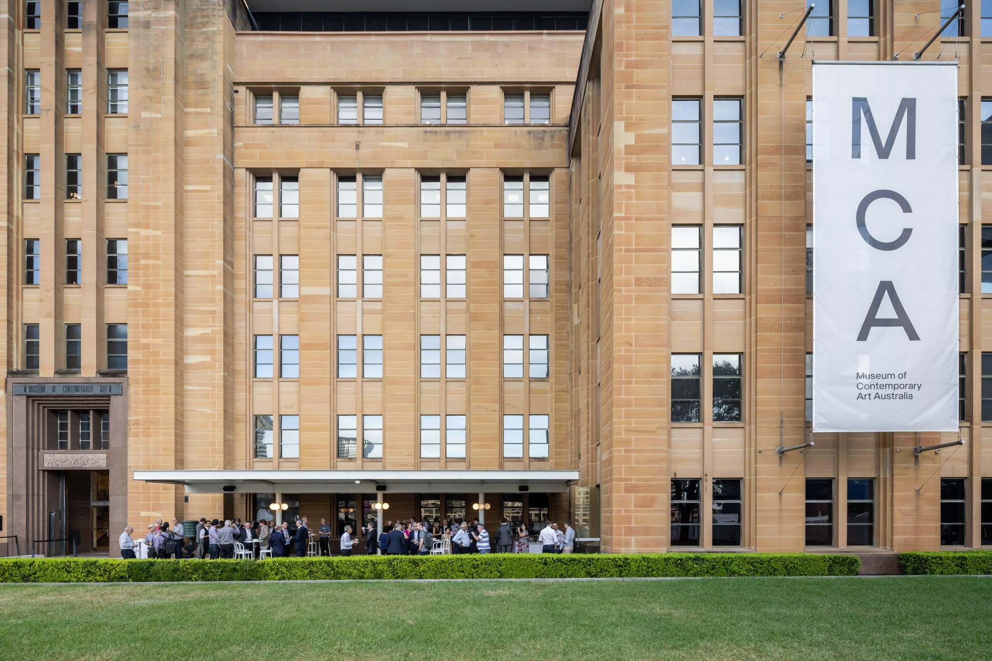 People gathered outside the Museum of Contemporary Art Australia in front of a large beige building with many windows, and a large white vertical banner reading 'MCA'.