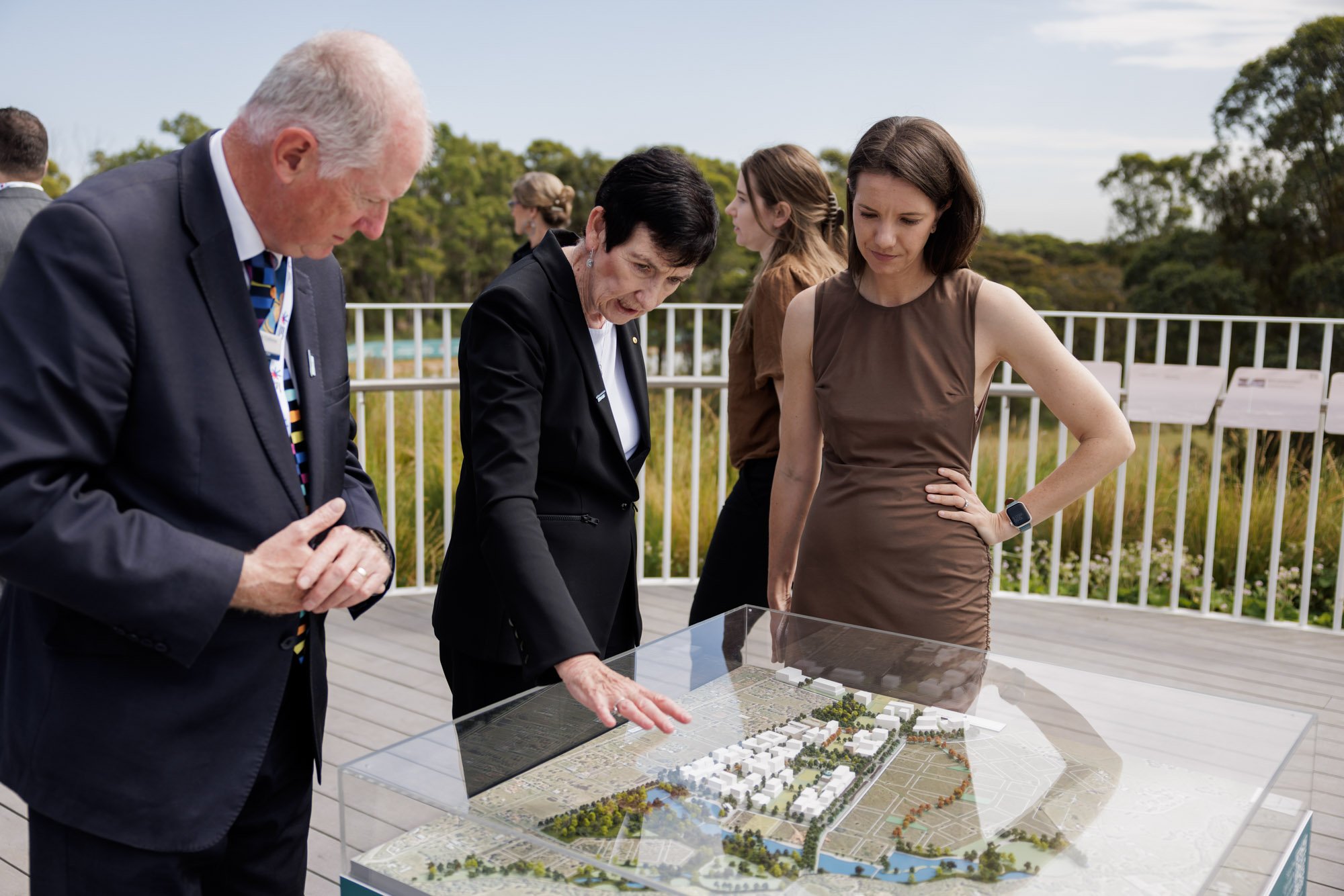 Four people are looking at a scale model of a planned development or park, outdoors on a deck with trees and sky in the background.
