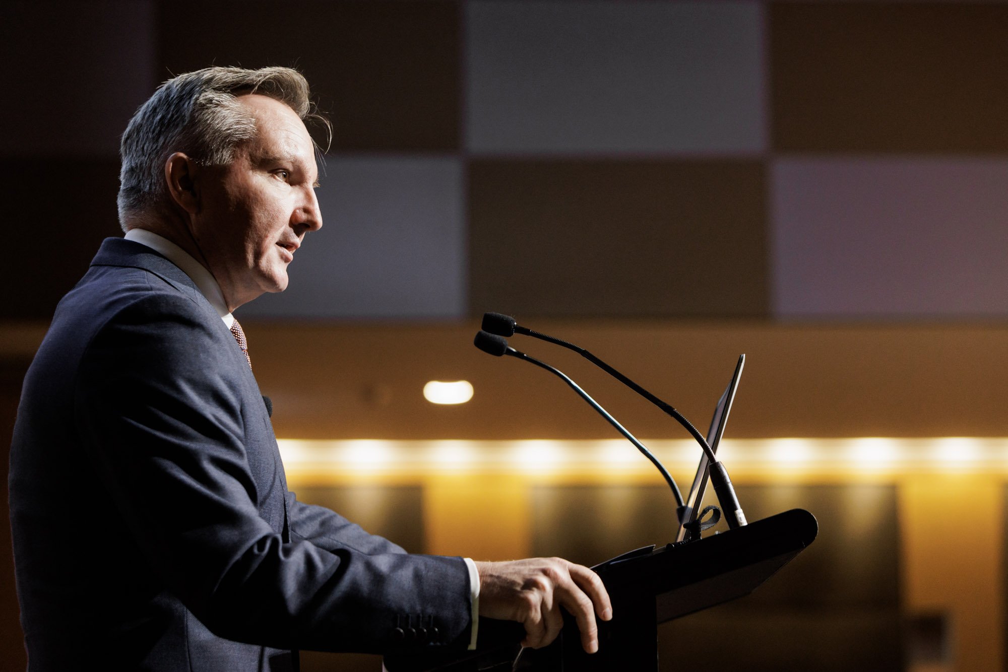 A man in a dark suit giving a speech at a podium with two microphones, in a dimly lit room with modern decor.