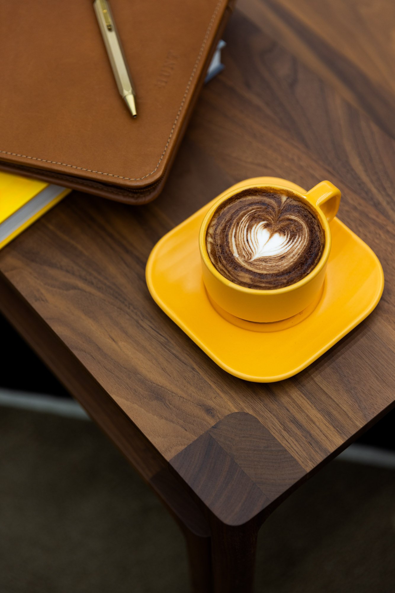 A yellow coffee cup with latte art on top, placed on a matching yellow saucer on a wooden table. Part of a brown leather notebook with a pen on top is visible next to the cup.
