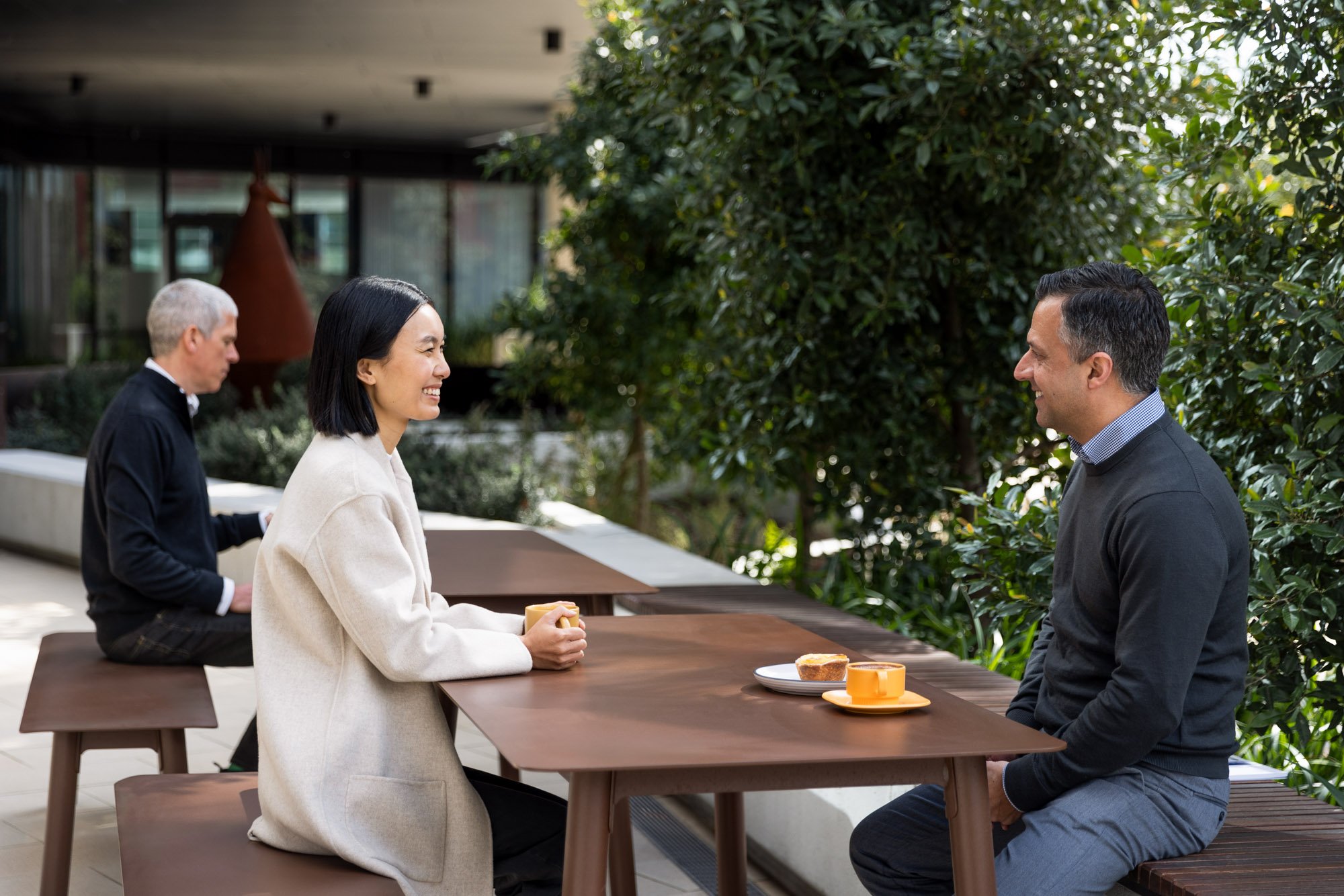 Two people sitting at a wooden table outdoors, smiling and facing each other. The woman has black hair, is wearing a beige coat, and holding a yellow mug. The man has dark hair, is wearing a dark sweater over a collared shirt, and has a plate with a 