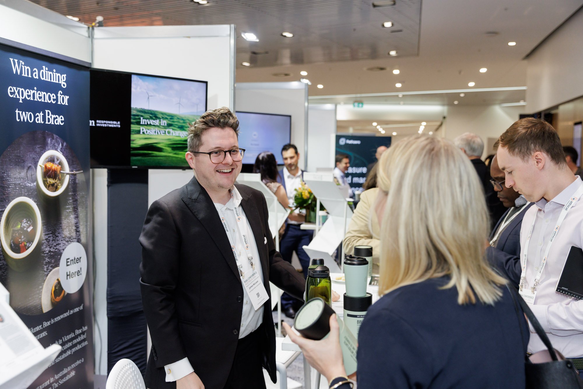 A man in a black suit and glasses smiling at a booth at a conference or trade show, speaking with a woman with blonde hair, while other attendees look on or browse displays in the background.