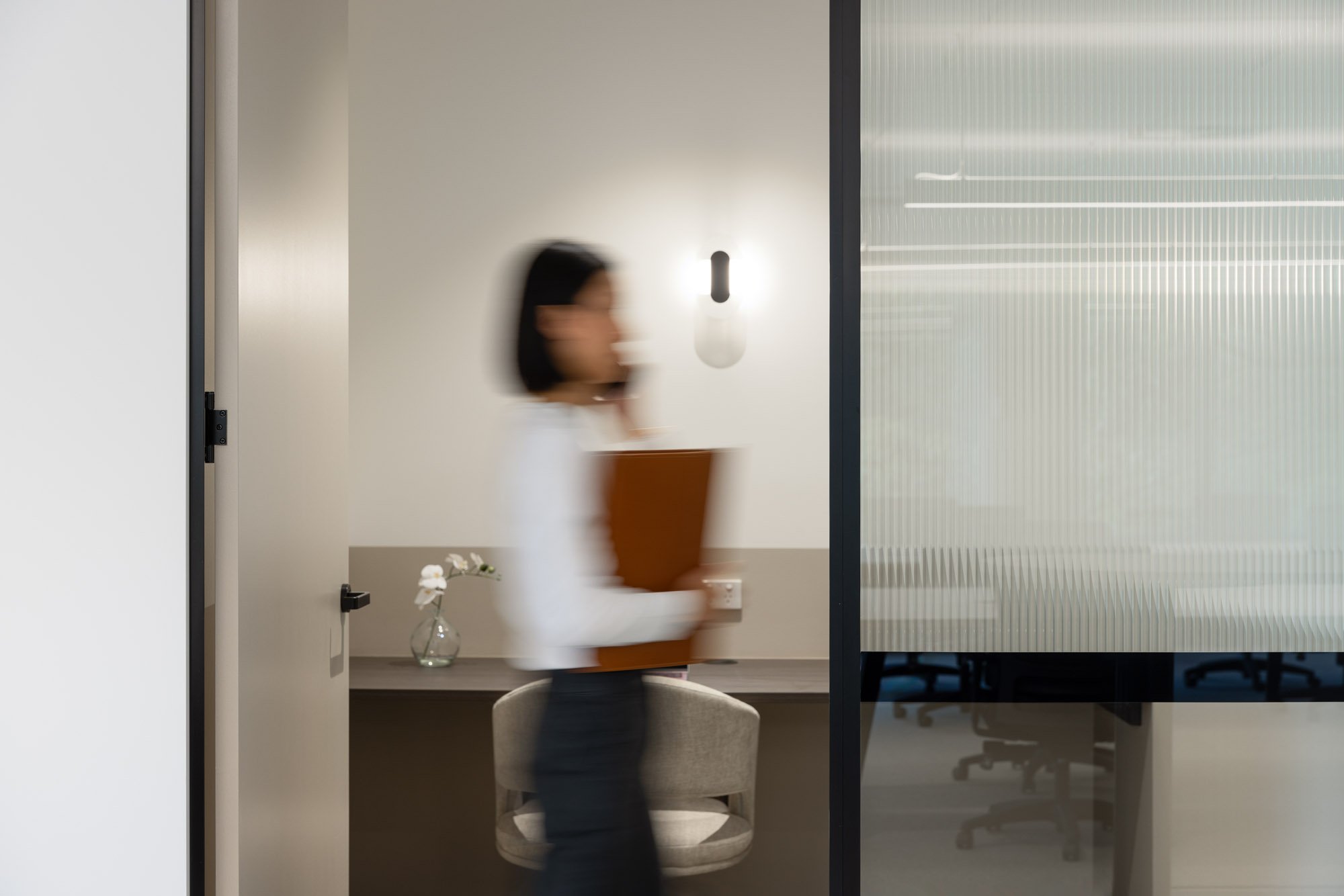 A blurred woman walking past an office cubicle with a chair and a small vase of flowers, holding a folder while talking on the phone inside a modern office space.