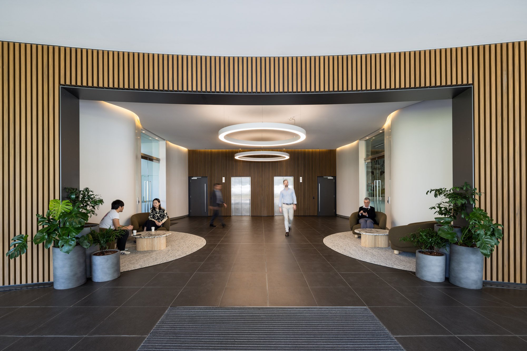 Modern hotel lobby with wooden paneling, circular ceiling lights, seating areas with chairs and potted plants, and two elevators in the background.