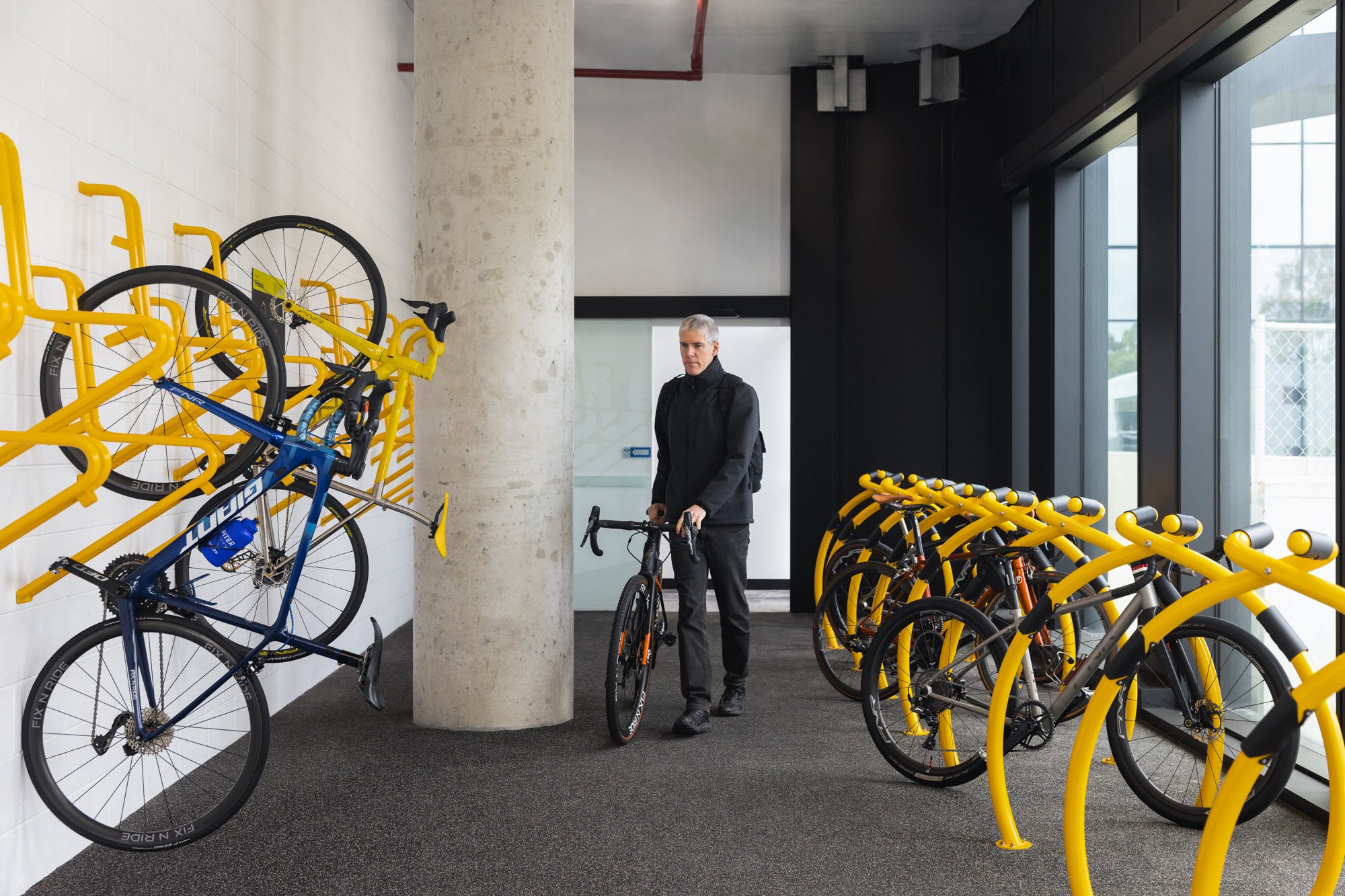 A man with gray hair, dressed in black, standing next to a black bicycle inside a bike rental area. The area has yellow bike racks on the right and a wall-mounted bike rack on the left with yellow and blue bicycles. The space features large windows a