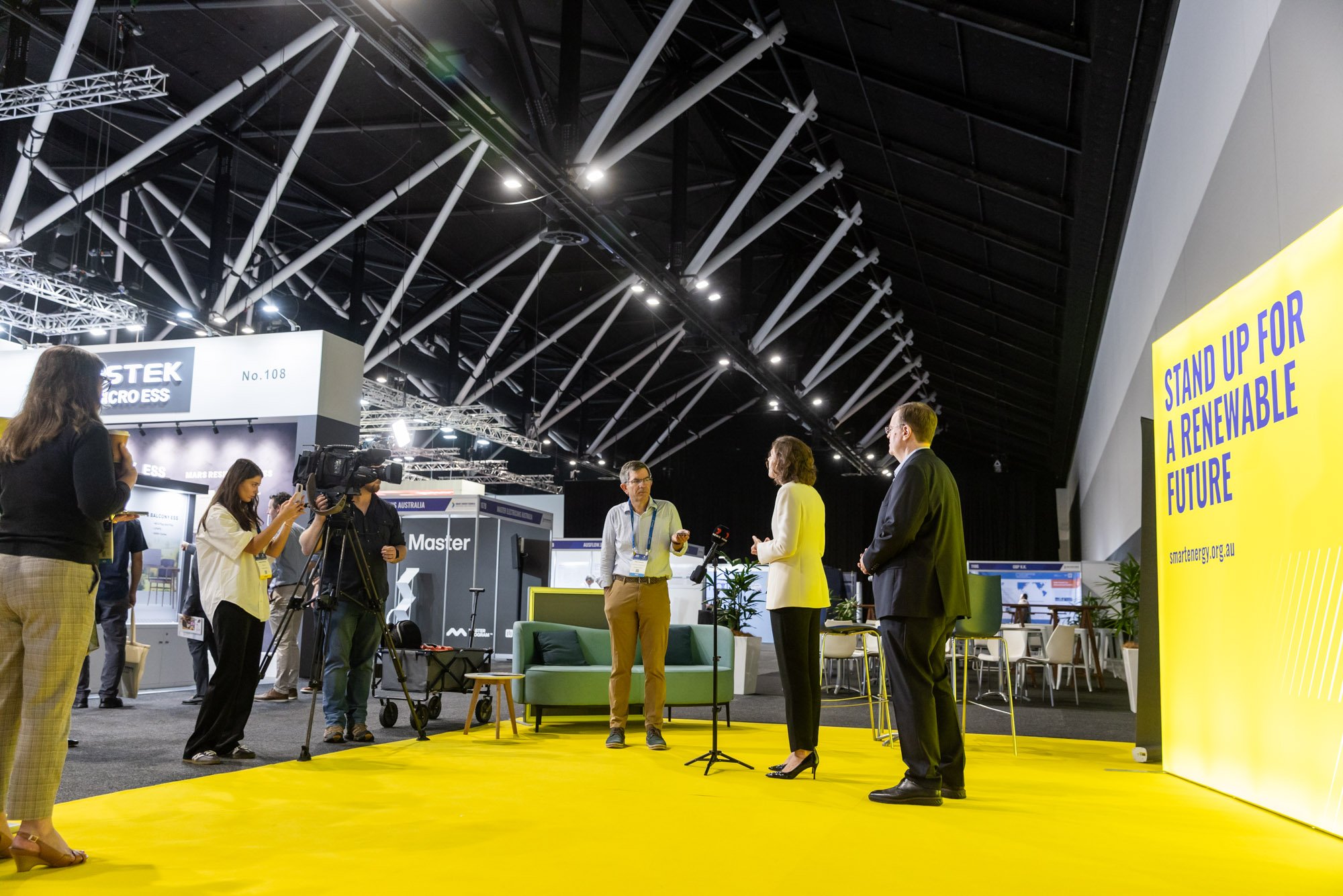 People on a yellow stage at an event or conference, with a large yellow sign that says "STAND UP FOR A RENEWABLE FUTURE" on the right. Several individuals are being interviewed or recorded, with cameras and microphones present, inside a large exhibit