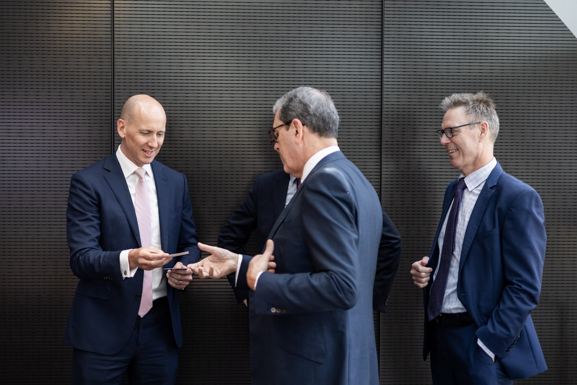 Four businessmen in formal suits standing and talking in front of a dark textured wall.