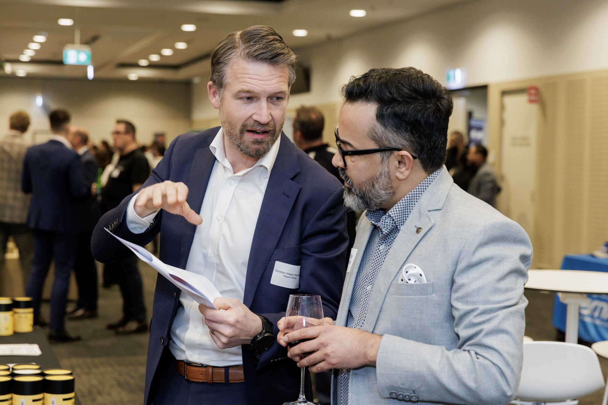 Two professional men engaged in conversation at a networking event, one holding a glass of wine, in a conference room with other attendees in the background.