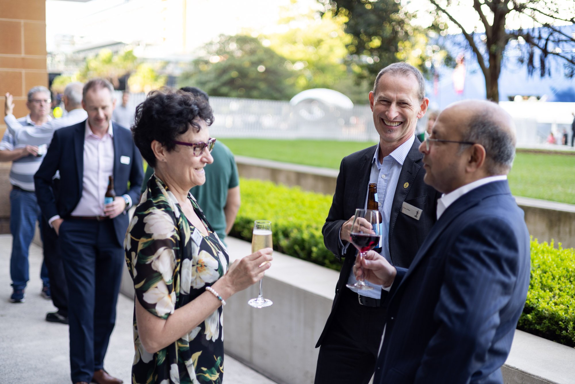 Group of professionally dressed people at an outdoor social event, holding drinks and engaging in conversation, with a green park and trees in the background.