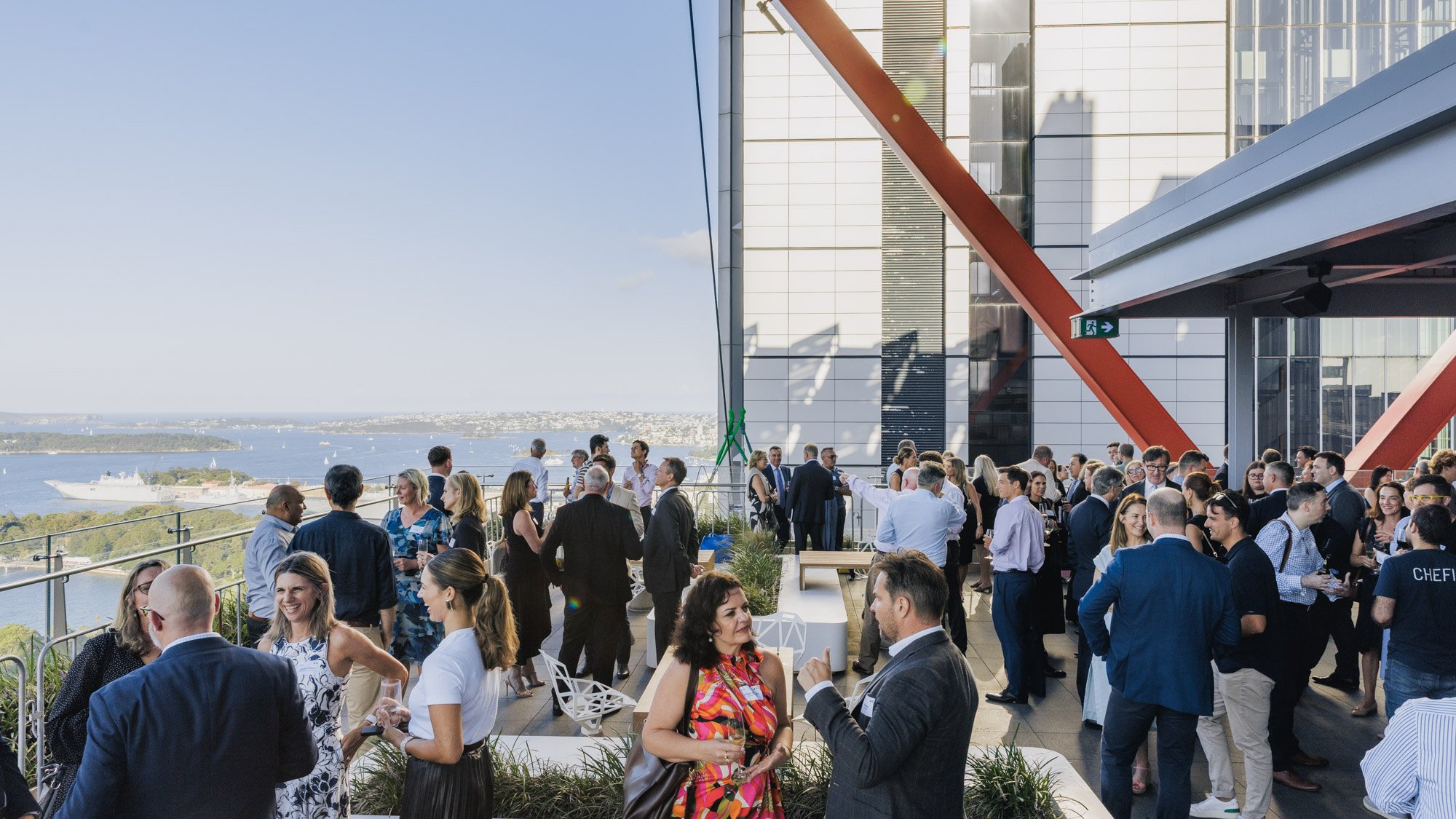 Many people attending a social event on a rooftop terrace overlooking a river and city skyline, with modern glass buildings and a clear blue sky.