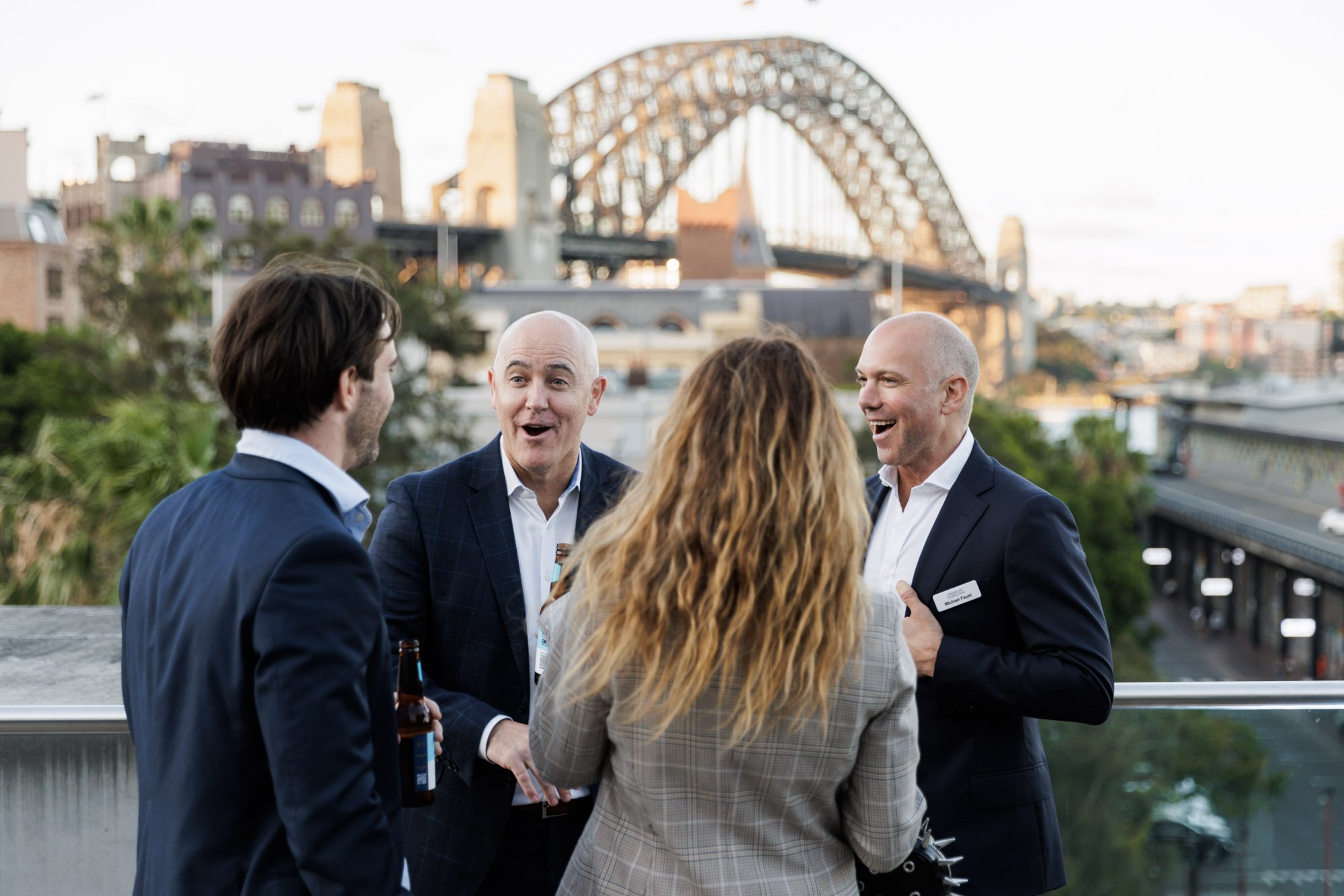 Four people, two men and one woman, talking at a social event outdoors with the Sydney Harbour Bridge in the background.