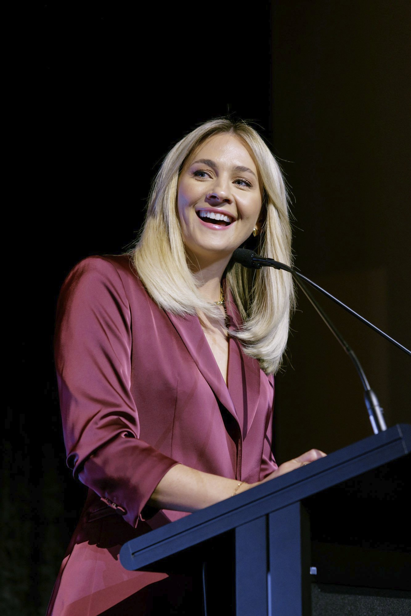 A woman with blond hair, wearing a burgundy satin blazer, is speaking at a podium with a microphone at an event. She is smiling.