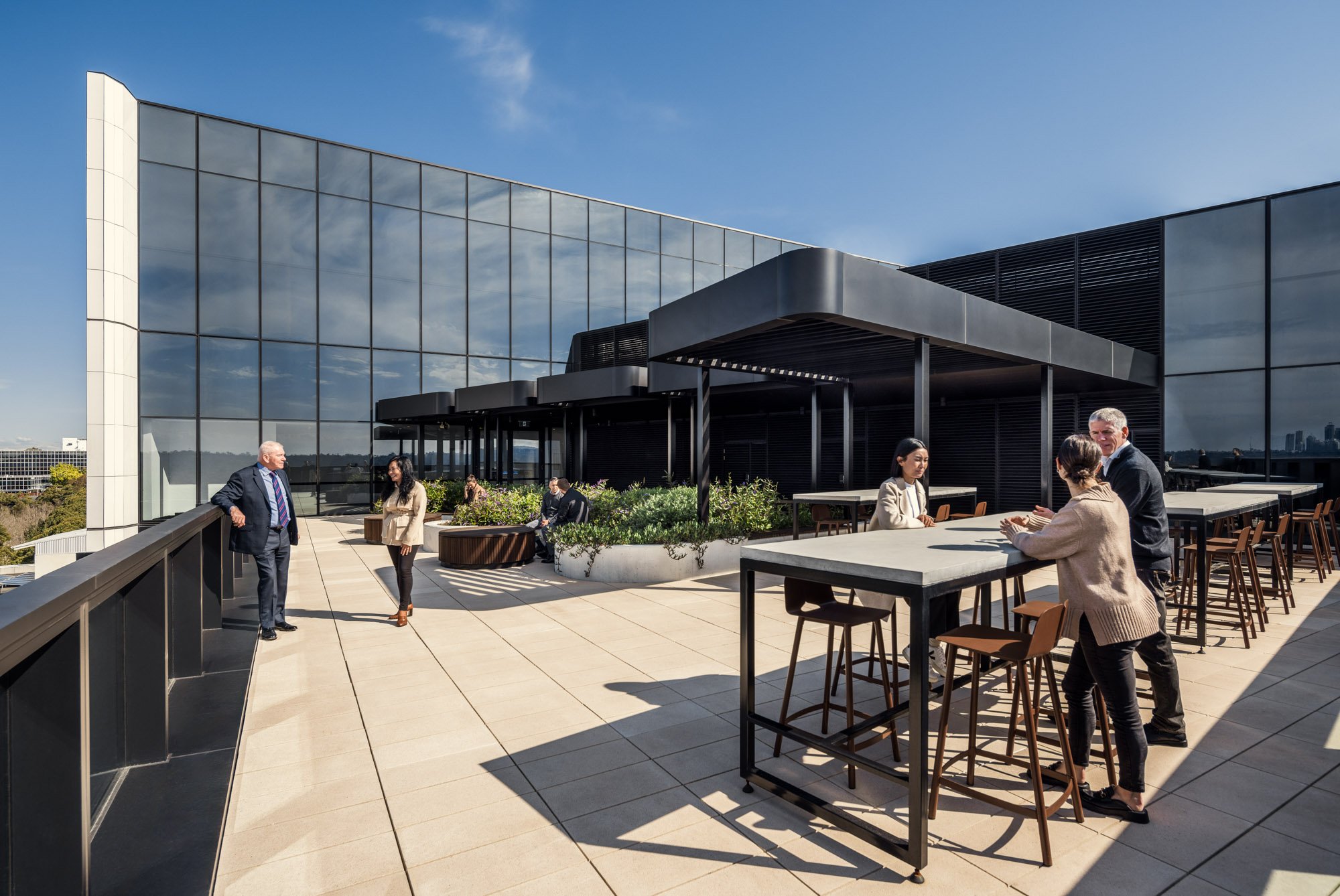 People on a rooftop terrace with modern glass building, some sitting, some standing, under clear sky.