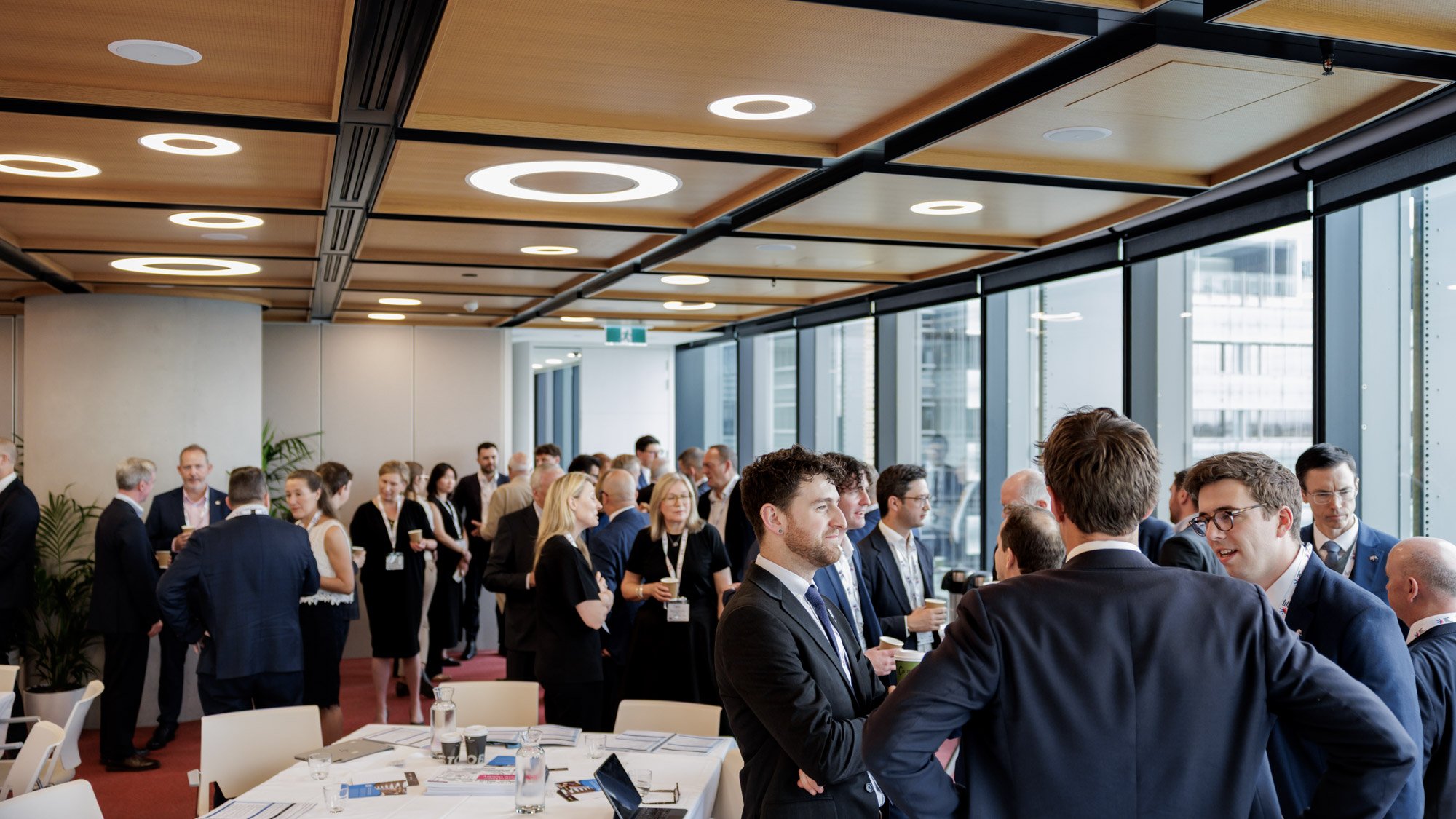Business conference or networking event with professionals in suits socializing in a modern, high-rise office meeting room with large windows and city view.