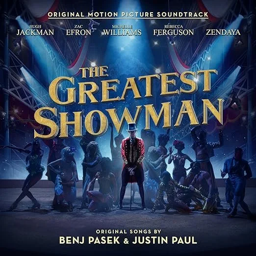 Album cover of The Greatest Showman: Original Motion Picture Soundtrack featuring the film’s title in bold gold lettering on a dark blue circus stage background with spotlights shining, and silhouettes of performers in dynamic poses beneath the title