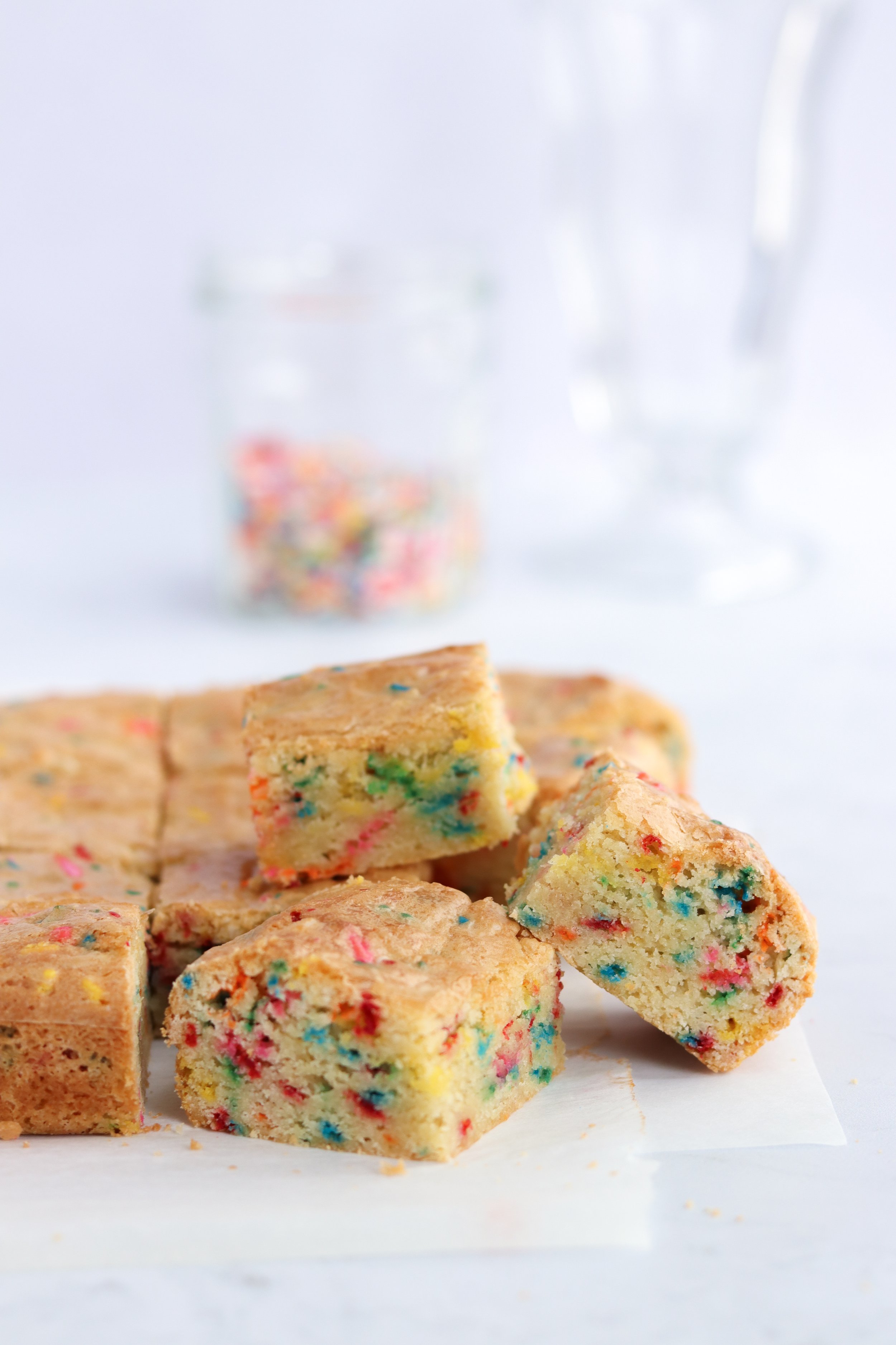 confetti cookie blondies, funfetti cookie blondies, cookie bars, cut into squares, gluten free, dairy free, soy free, white parchment paper, jar of sprinkles, glass