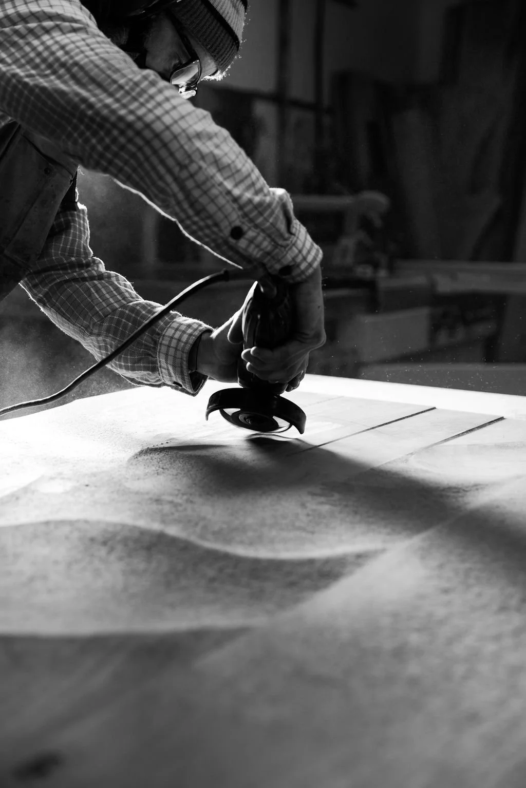 Lars wearing glasses, a beanie, and a plaid shirt is using a handheld angle grinder on a piece of wood in a workshop.