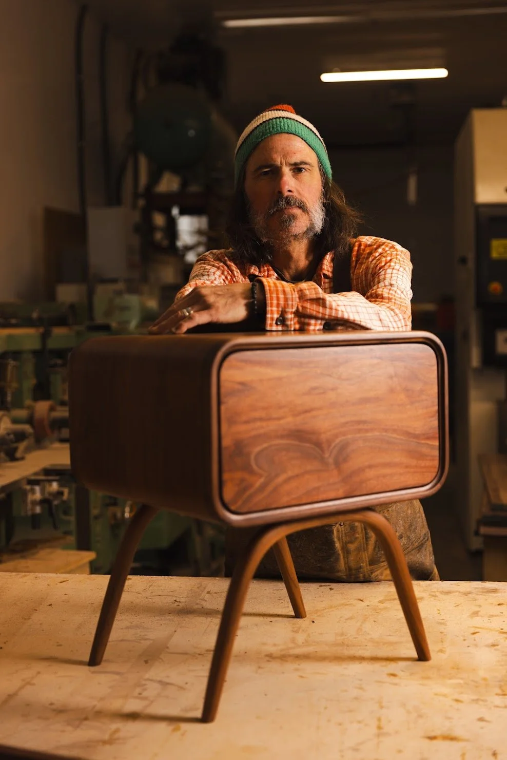 Lars wearing a striped beanie and plaid shirt, standing behind a retro wooden radio with long tapered legs in a workshop setting.