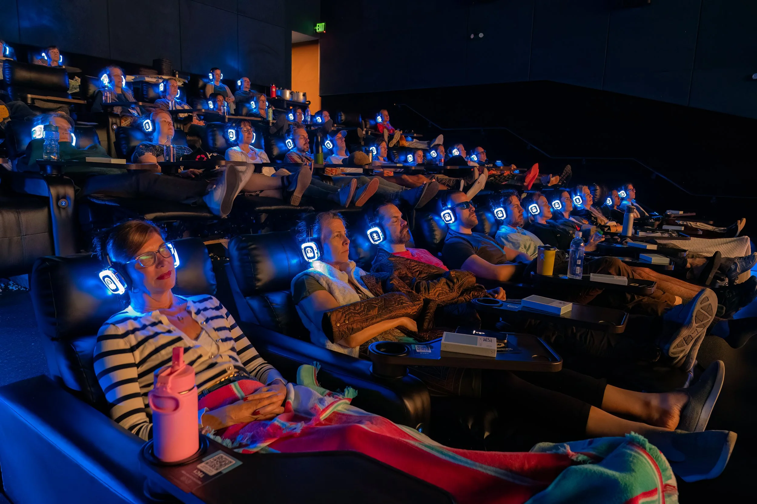 People sitting in recliner chairs in a dark theater, wearing headphones, watching a presentation or movie.