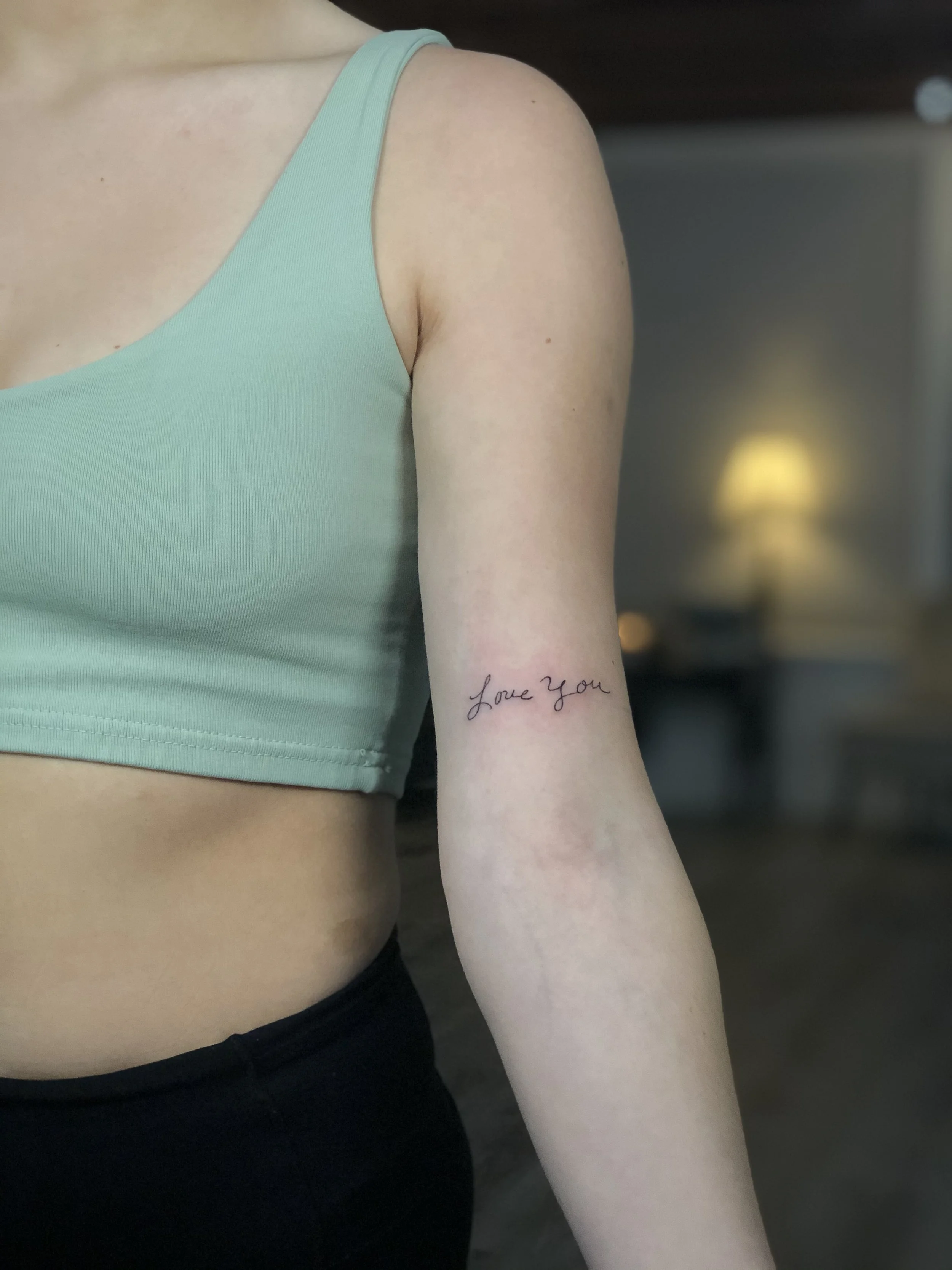 Close-up of a person's upper body, focusing on their arm and side, with a tattoo that says "Love You" on their forearm, wearing a light turquoise tank top and black pants.