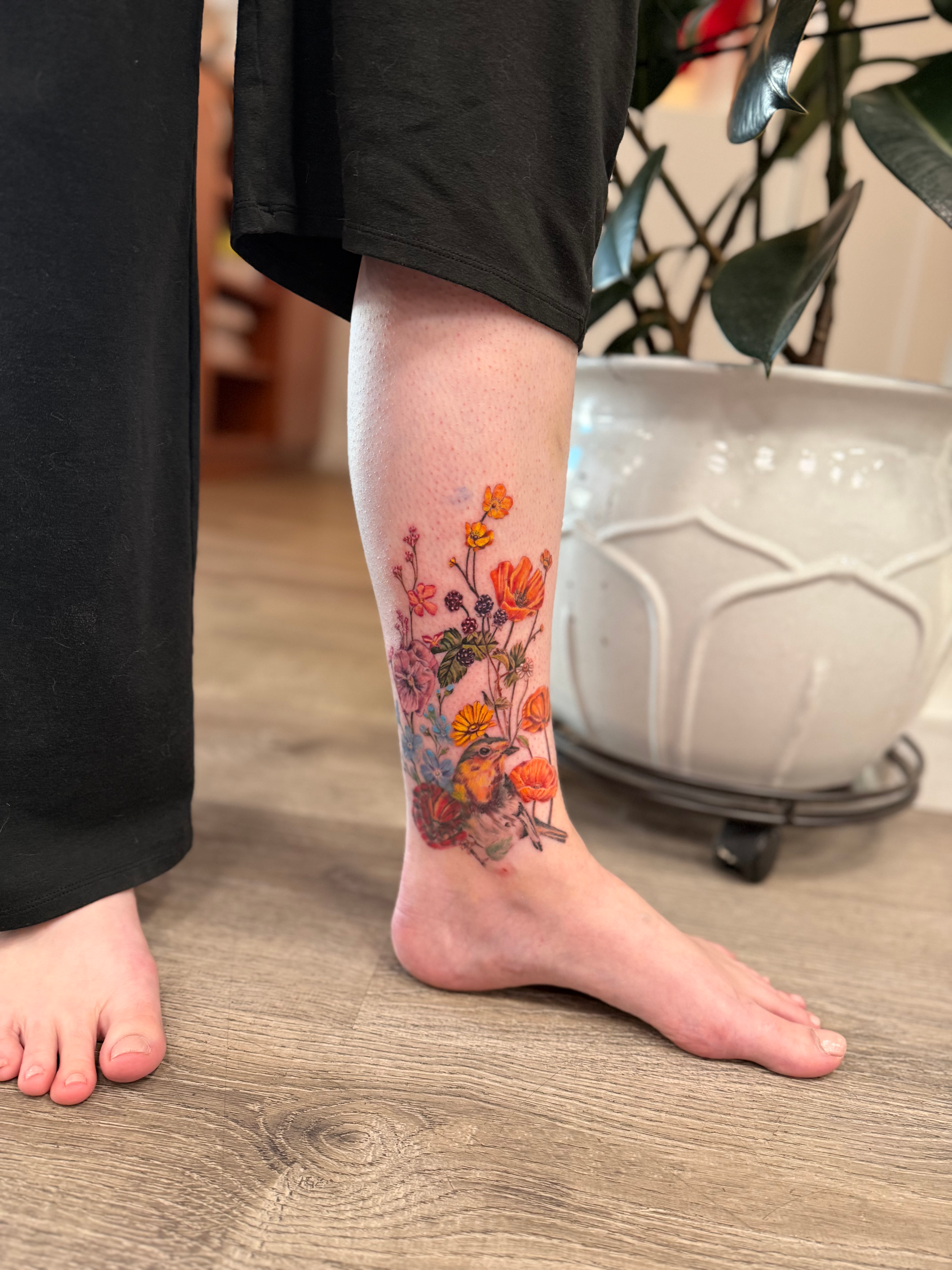 A person's lower leg and foot with a colorful floral cover-up tattoo. The person is wearing a black garment, and the background includes a wooden floor, a potted plant.