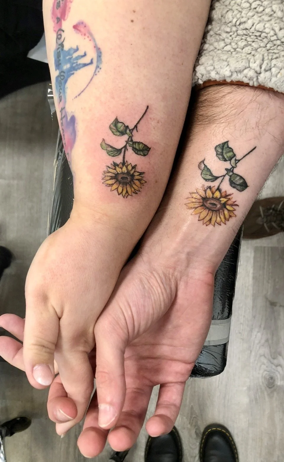 Matching tattoos of a sunflower with a stem and leaves is visible on a person's lower forearm, just above the wrist.
