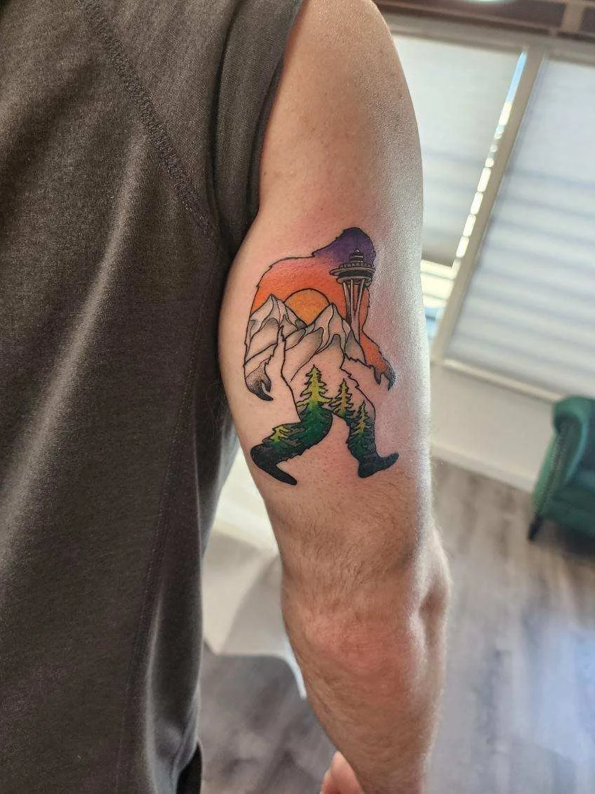 Tattoo of a mountain landscape with trees, a river, and the Space Needle against a sunset or sunrise sky.