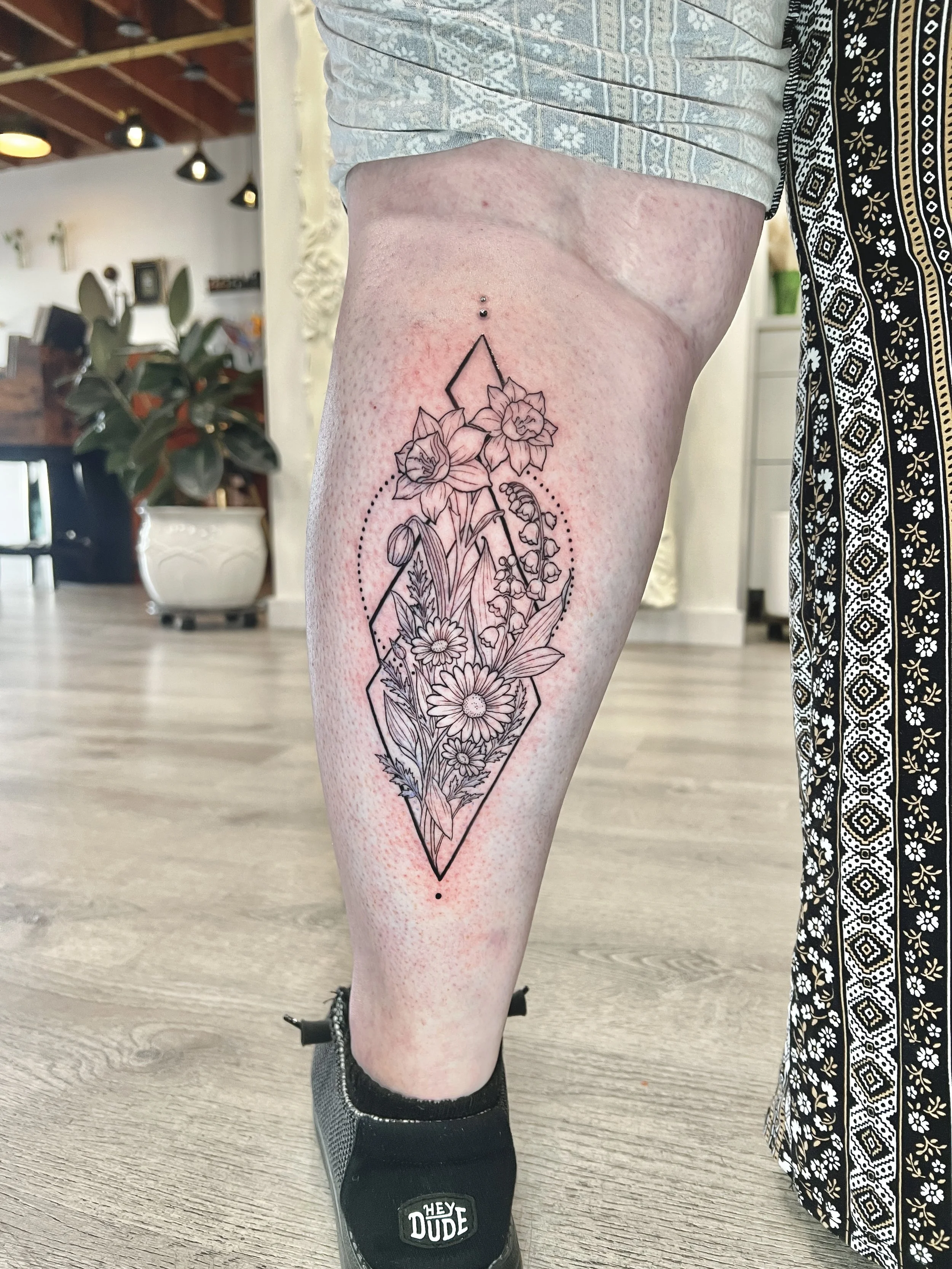 A person's leg with a fine-line floral tattoo on back of leg. The tattoo features daffodils, lilies, and other flowers inside a geometric diamond shape. The person is wearing black sneakers with a white logo and a patterned skirt.