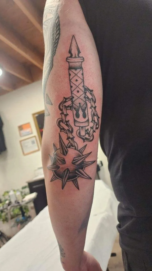 Tattoo of a spiked ball and chain with a dagger piercing through it on a person's forearm.
