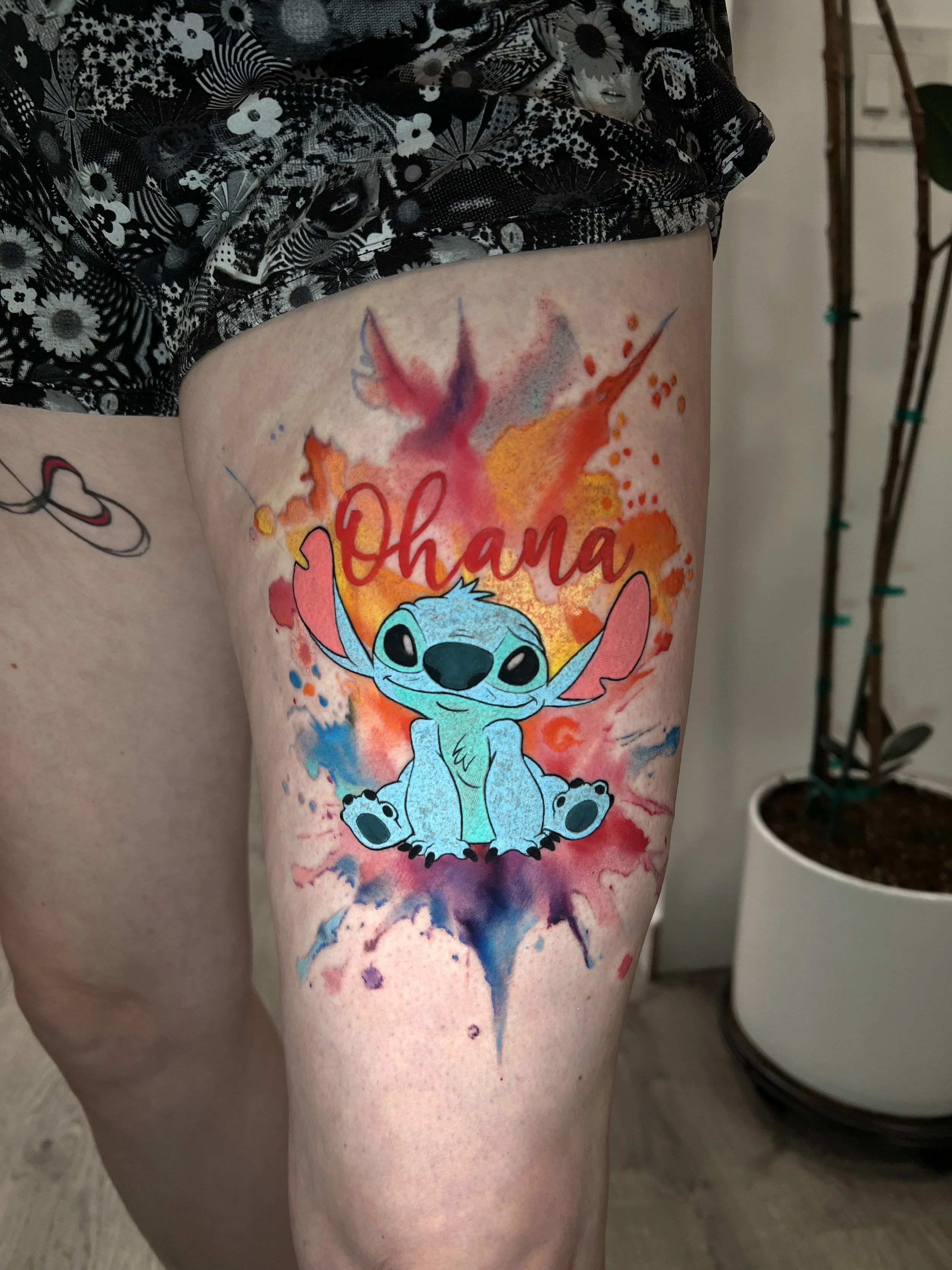 Colorful tattoo of Stitch from Lilo & Stitch with watercolor splash background and the name 'Ohana' on a person's thigh.