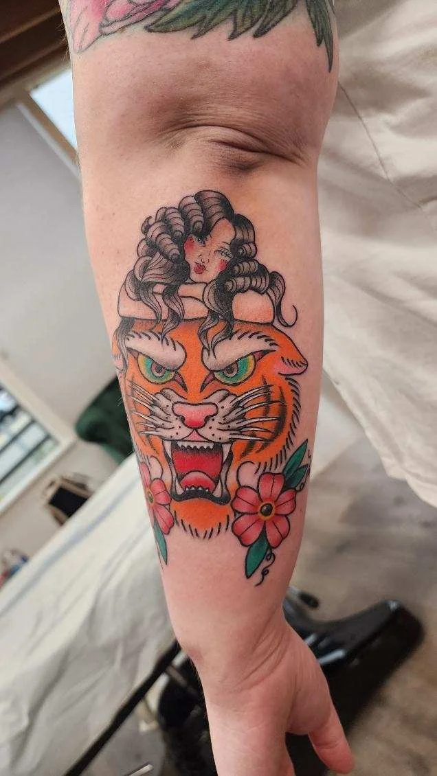 Tattoo of a woman with wavy hair resting her chin on a fierce, roaring tiger's head, surrounded by pink flowers with green leaves.