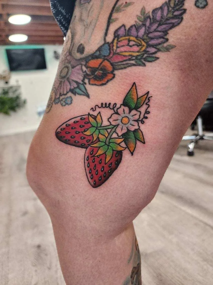 Tattoo of a strawberry with a small flower and leaves on a person's upper thigh.