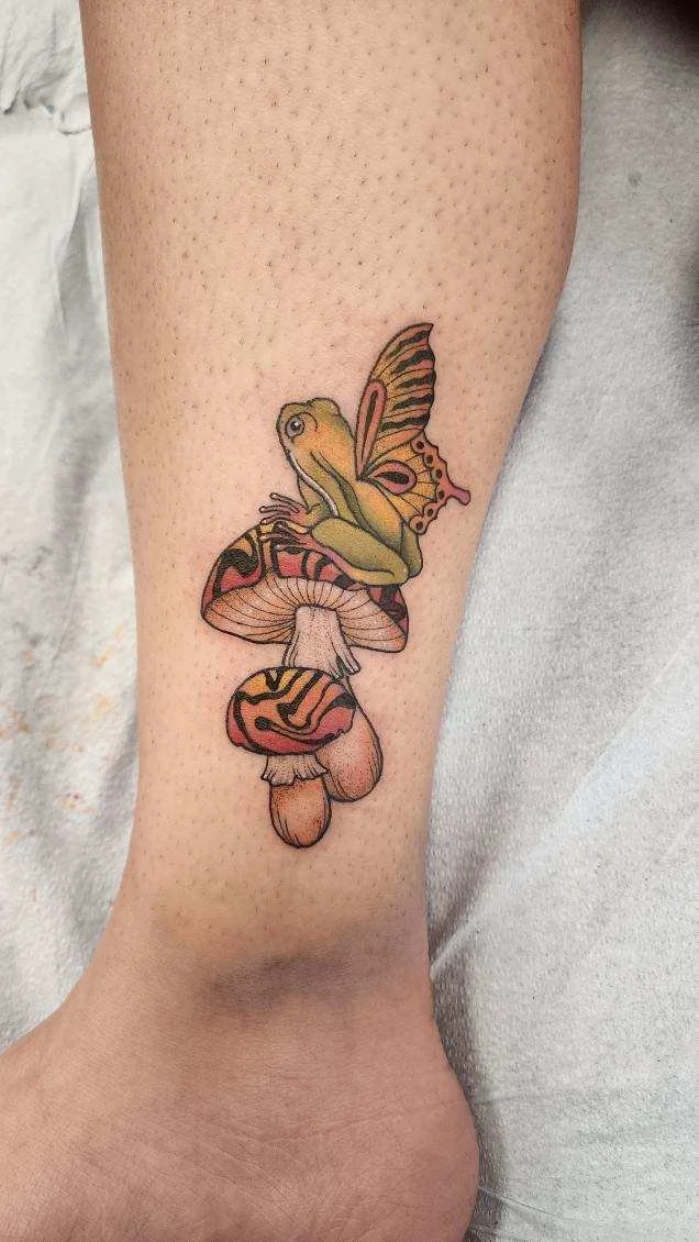 Tattoo of a frog with butterfly wings sitting on top of two mushrooms on a person's leg.