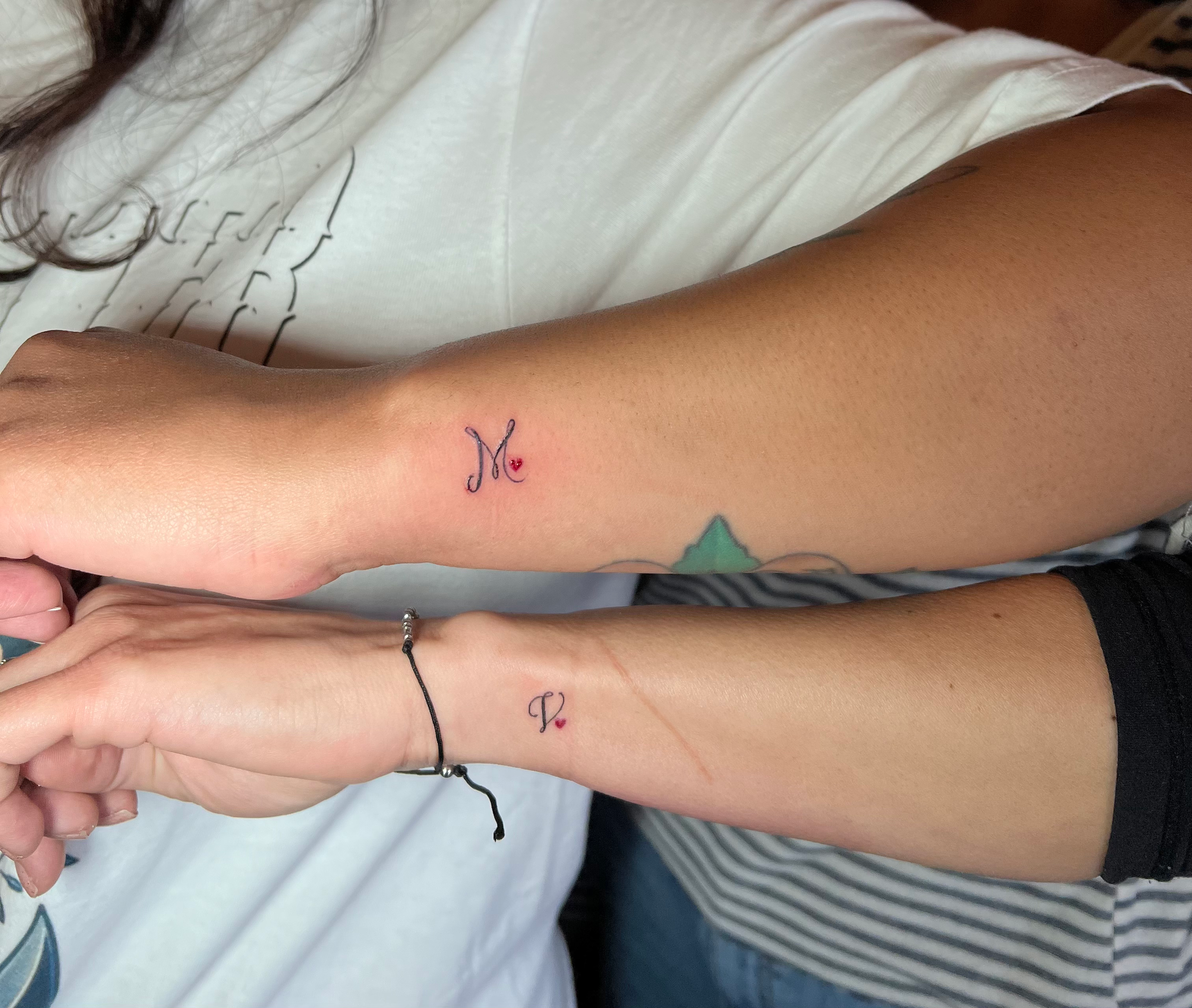 Close-up of matching tattoos showing a small tattoo of a heart with a letter 'M' and 'V' and a black string bracelet on their wrist. The background includes part of a white shirt and other indistinct objects.