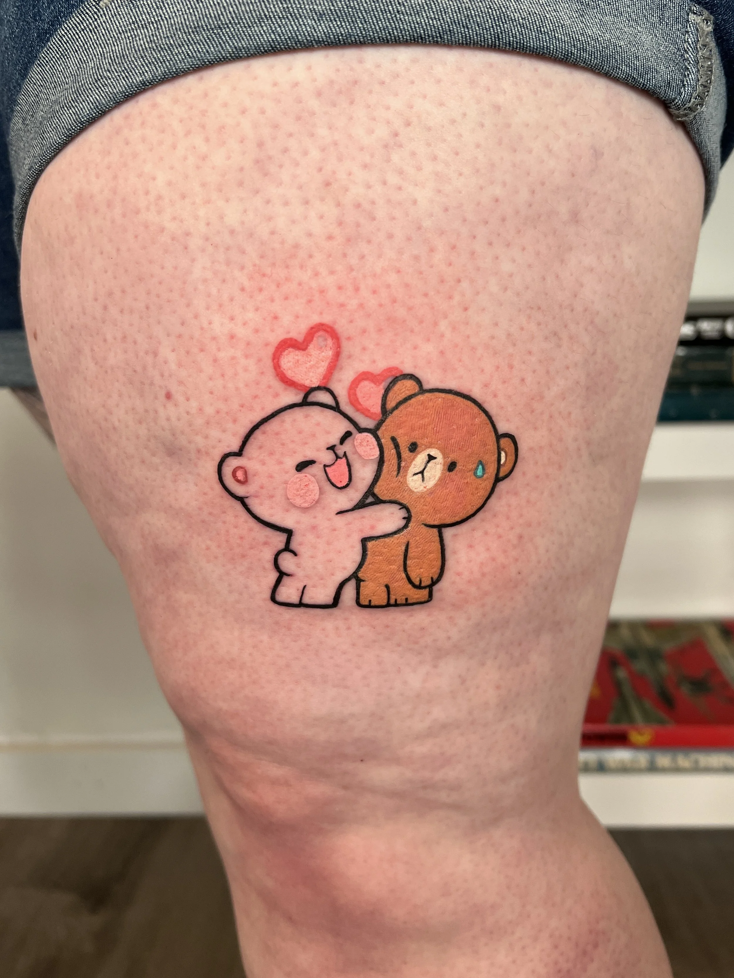 Tattoo of two cartoon bears, one pink and one brown, with pink and red hearts above them, on a person's thigh.