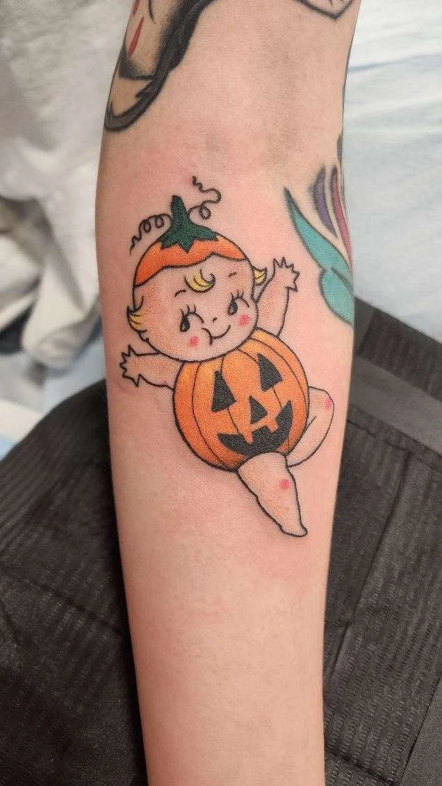 Tattoo of a cute baby with a pumpkin costume, featuring a Jack-O'-Lantern face, on a person's forearm.
