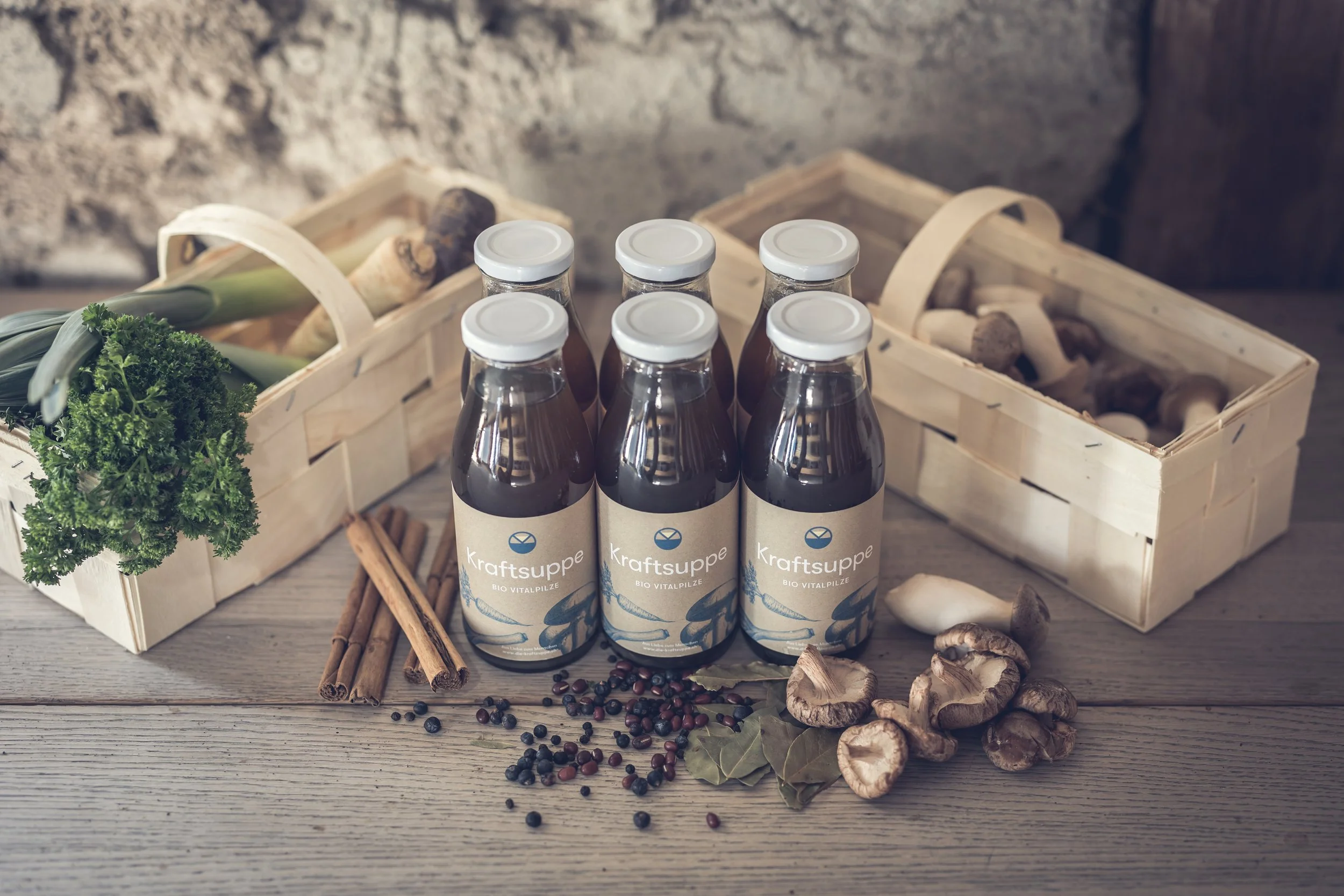 Flaschen Kraftsuppe auf einem Holztisch, umgeben von frischen Kräutern, Pilzen, Pfefferkörnern und Zimtstangen, mit einem Holzkasten im Hintergrund.