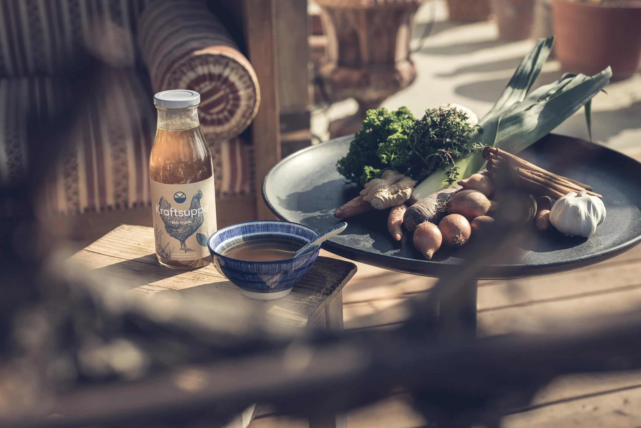 Ein Tisch mit Flasche Bio-Huhn Kraftsuppe, eine Schüssel mit Brühe, Knoblauch, Zwiebeln, Ingwer, frische Kräuter und Gemüse auf einer sonnigen Terrasse.
