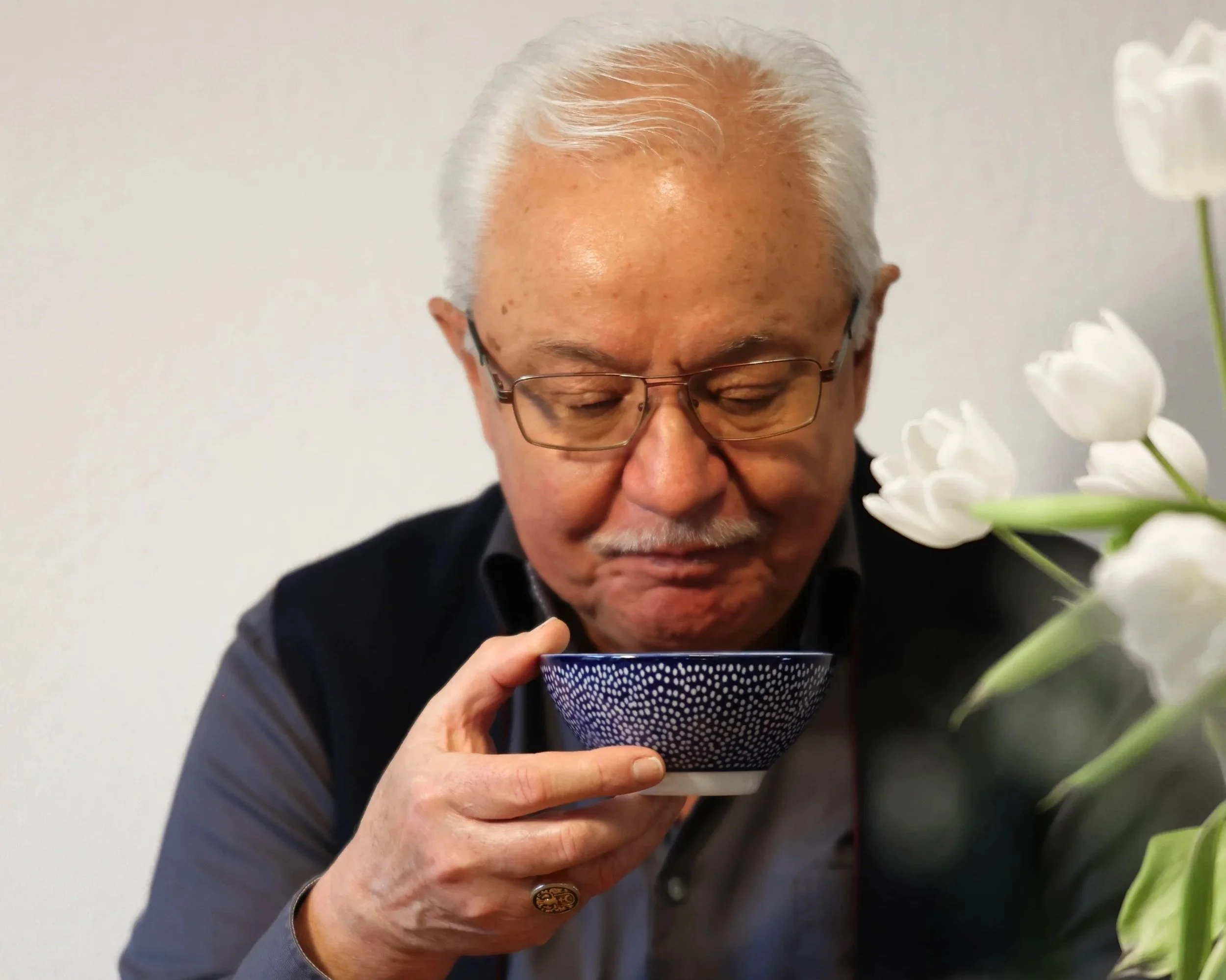 Ein älterer Mann mit weißen Haaren und Brille trinkt aus einer Schale Kraftsupe, während er neben weißen Blumen steht.