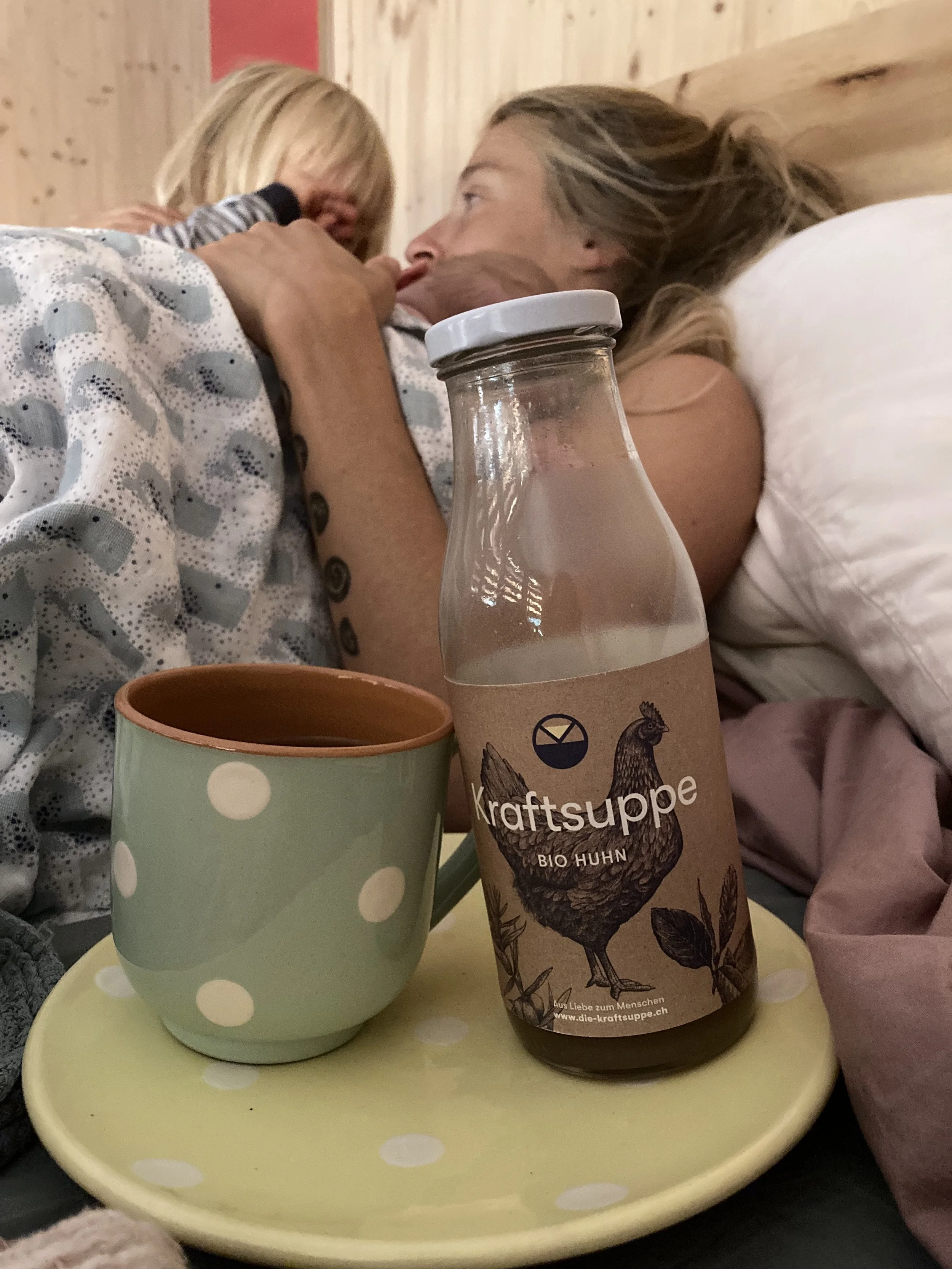 Eine Frau mit einem Baby im Wochenbett im Geburtshaus, während eine Flasche Kraftsuppe auf einem rosa Teller steht.