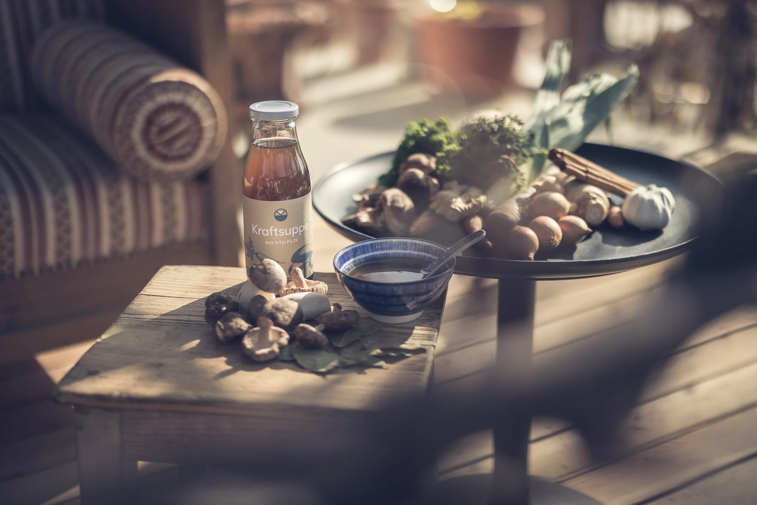 Frische Zutaten auf einem Holztisch, darunter Knoblauch, Ingwer, Zwiebeln, sowie ein Glas mit Bio Vitalpilz-Kraftsuppe, im Hintergrund eine Schale mit Kräutern.