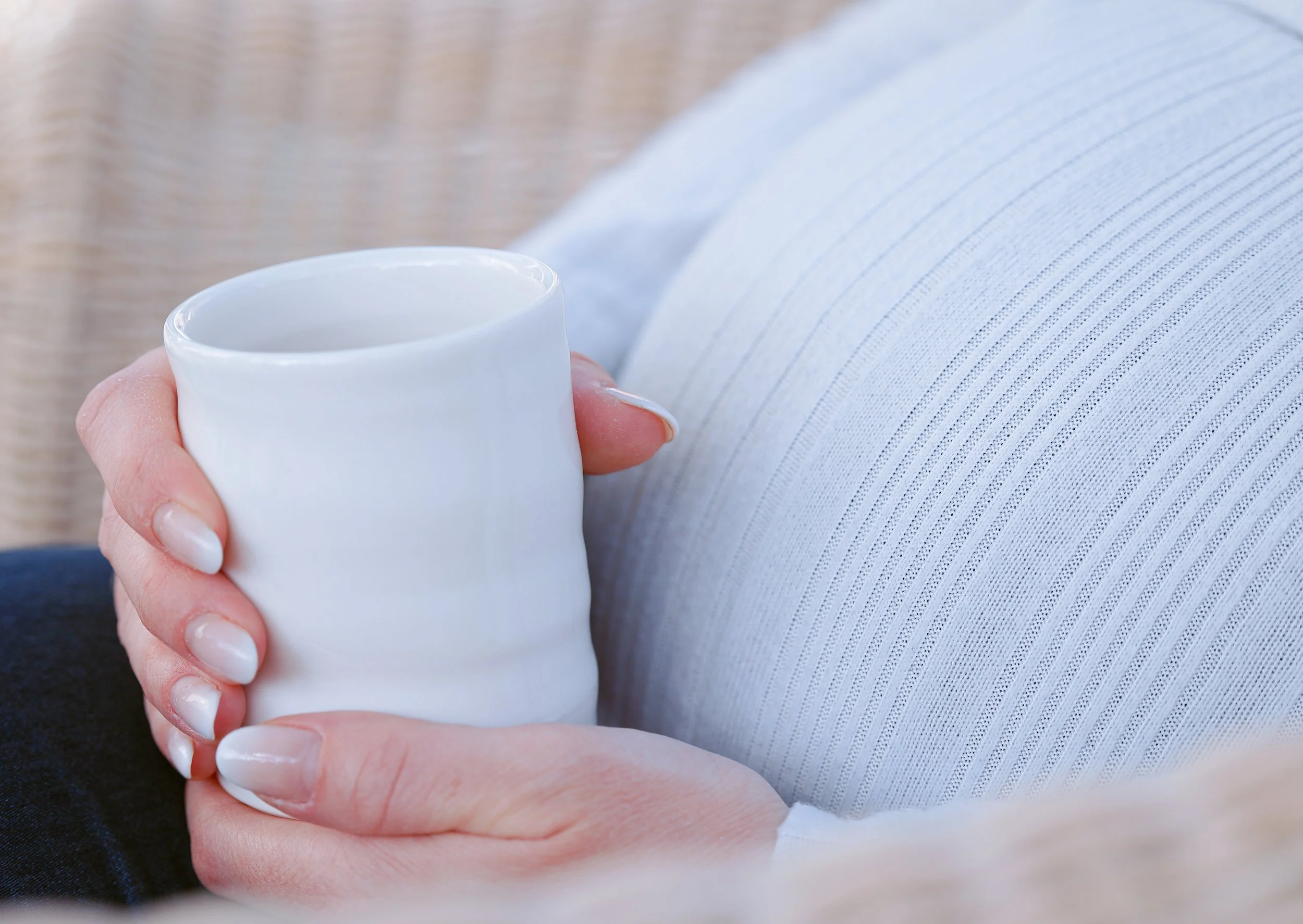 Eine schwangere Frau hält eine weiße Tasse mit Kraftsuppe in der Hand, während sie auf einem weißen Kleid oder Hemd sitzt.