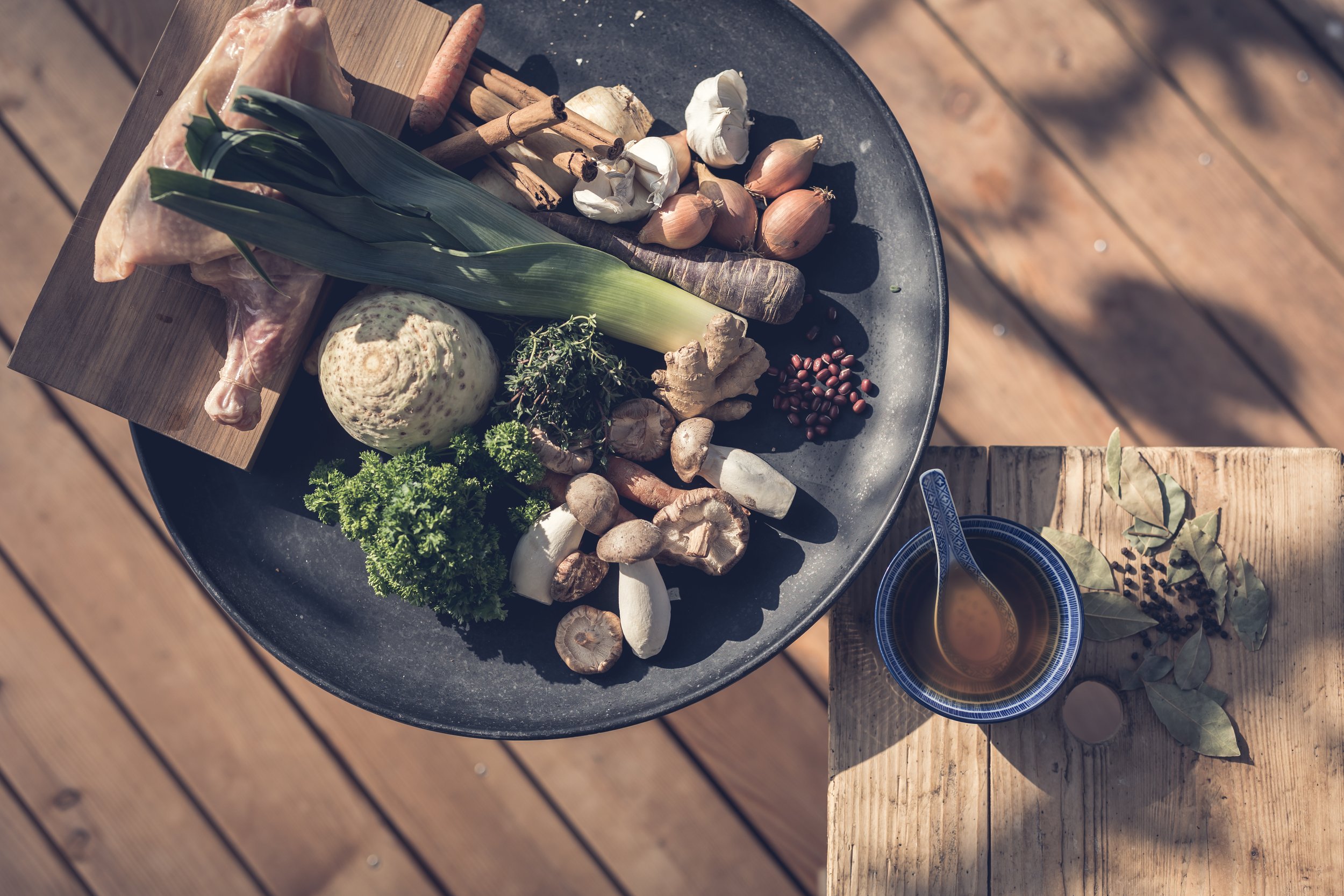 Verschiedene frische Zutaten auf einem schwarzen Teller, darunter Zwiebeln, Knoblauch, Ingwer, Petersilie, ein Lauch, Pilze, Wurzeln, weißes Gemüse und pflanzliche Gewürze, mit einer kleinen Schüssel mit Kraftsupp neben dem Teller auf einem Holztisch.