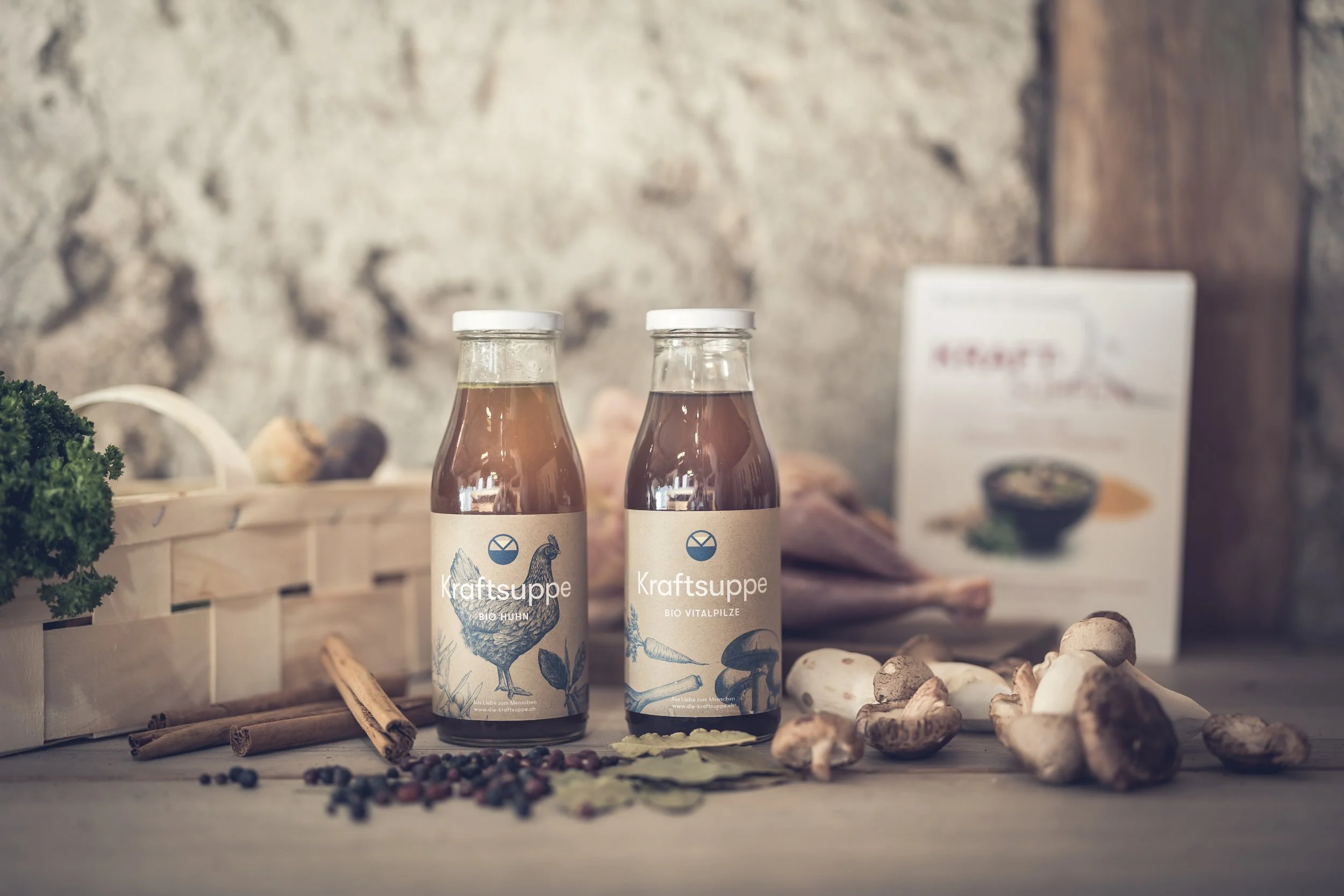 zwei Flaschen mit Kraftsuppe, Bio Huhn und Bio Vitalpilze, auf einem Holztisch, umgeben von Kräutern, Pilzen und Gewürzen, im Hintergrund ein Lebensmittelregal.