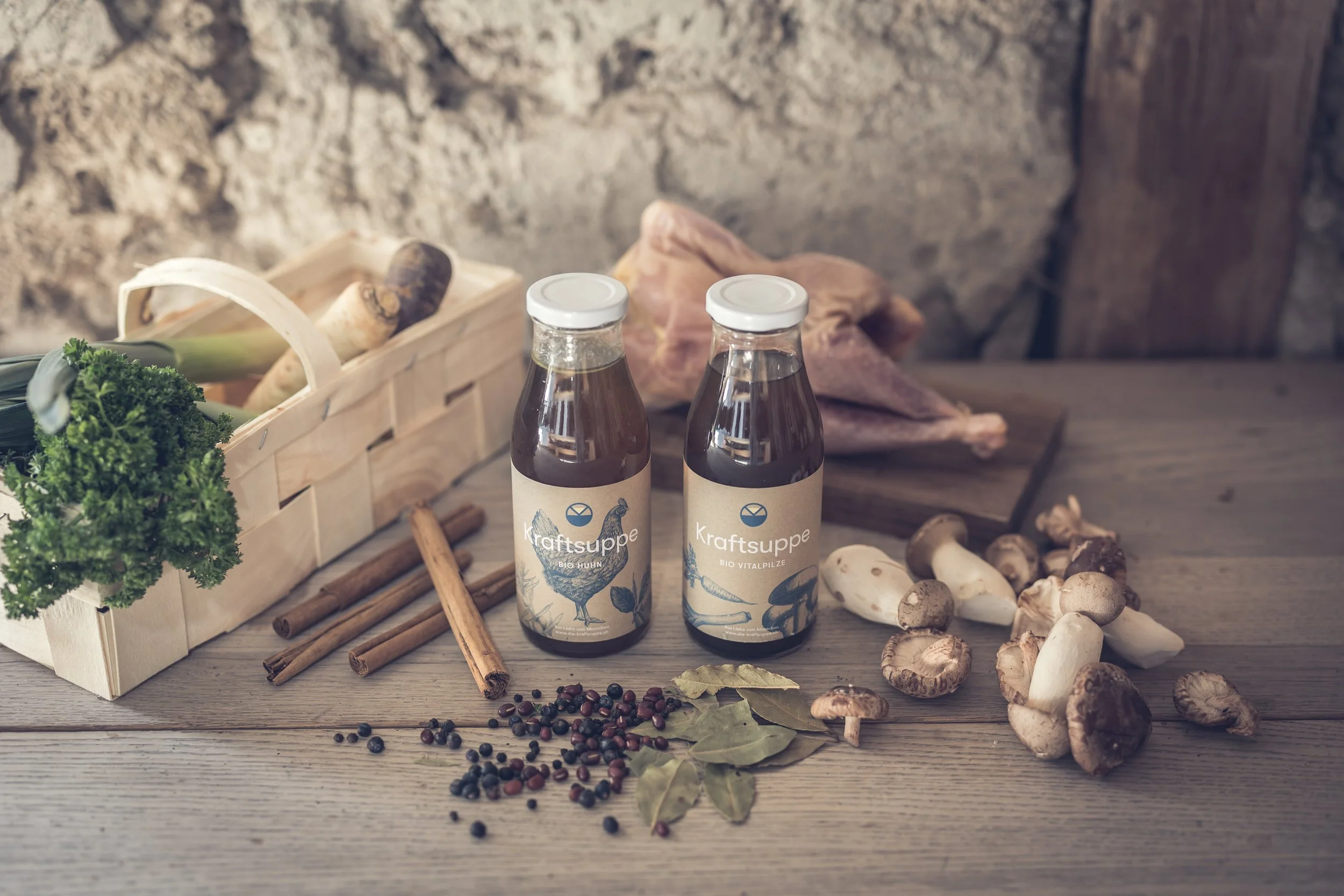 Zwei Flaschen Kraftsuppe mit Bio-Vitalpilzen und Bio-Huhn zeigen, stehen auf einem Holztisch. Im Hintergrund befinden sich frischer Ingwer, Pilze, getrocknete Kräuter, Pfefferkörner und andere Gewürze sowie eine Holzbox mit frischen Kräutern.